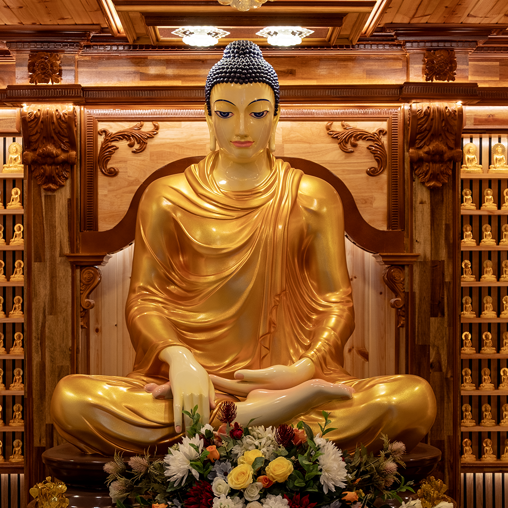 Golden Buddha statue in a wooden temple, surrounded by small Buddha statues on shelves, with a flower arrangement in front.