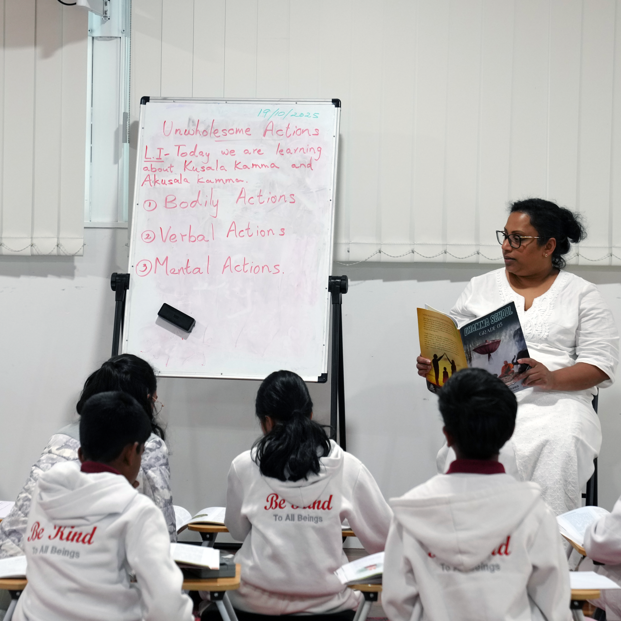 A teacher reading a book to students in a classroom, with a whiteboard in the background displaying lesson topics about actions and Kusaula Kamma.