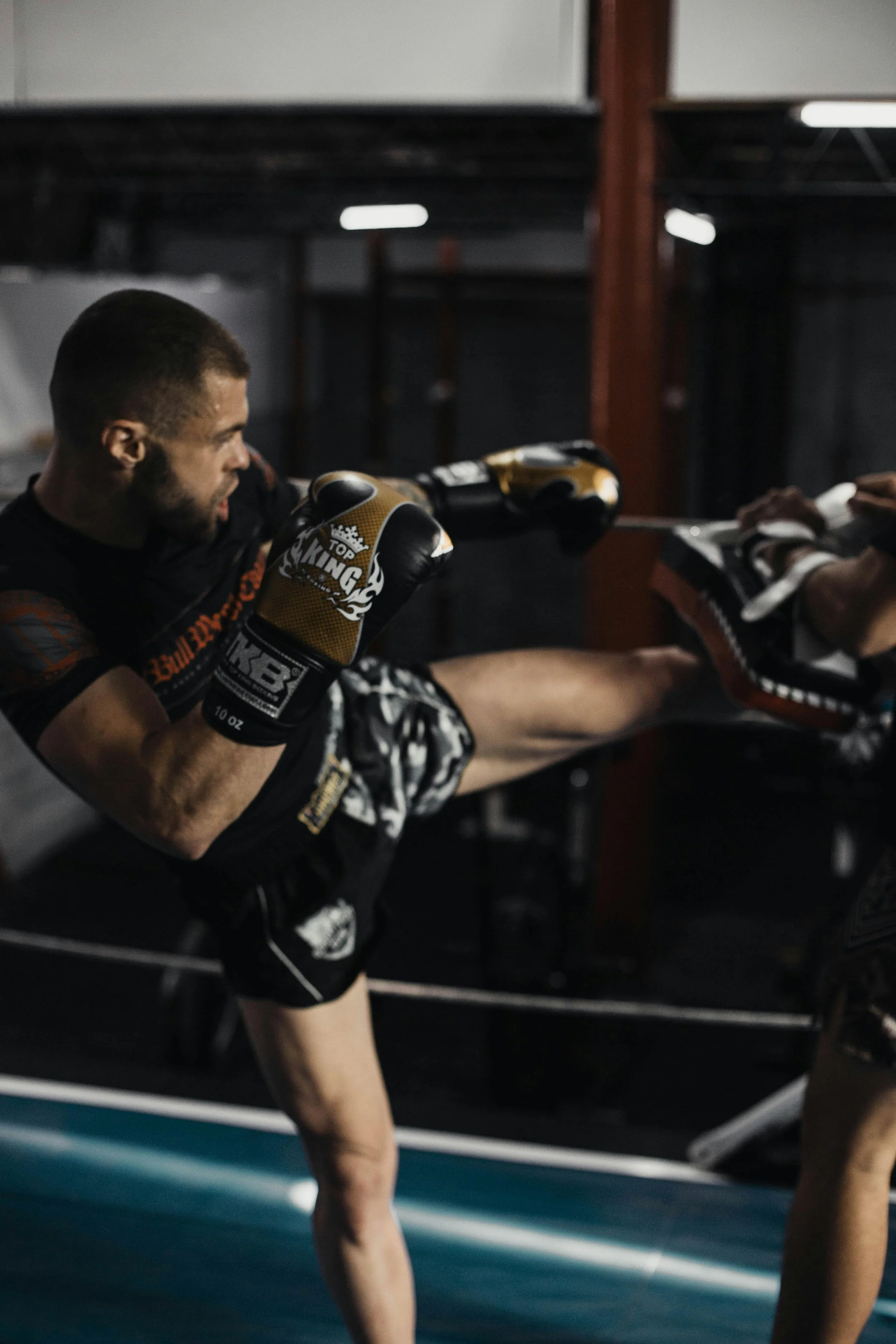 Vyras atlieka Kikbokso aukštą spyrį į pagalvę, kurią laiko kitas žmogus