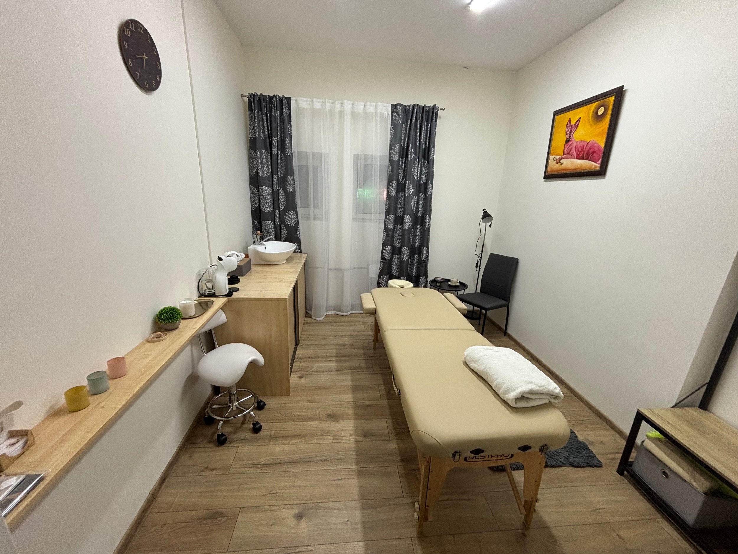 Masažo kambarys su masažo stalu, kėde, darbo stalu, lentyna su žvakėmis ir paveikslu