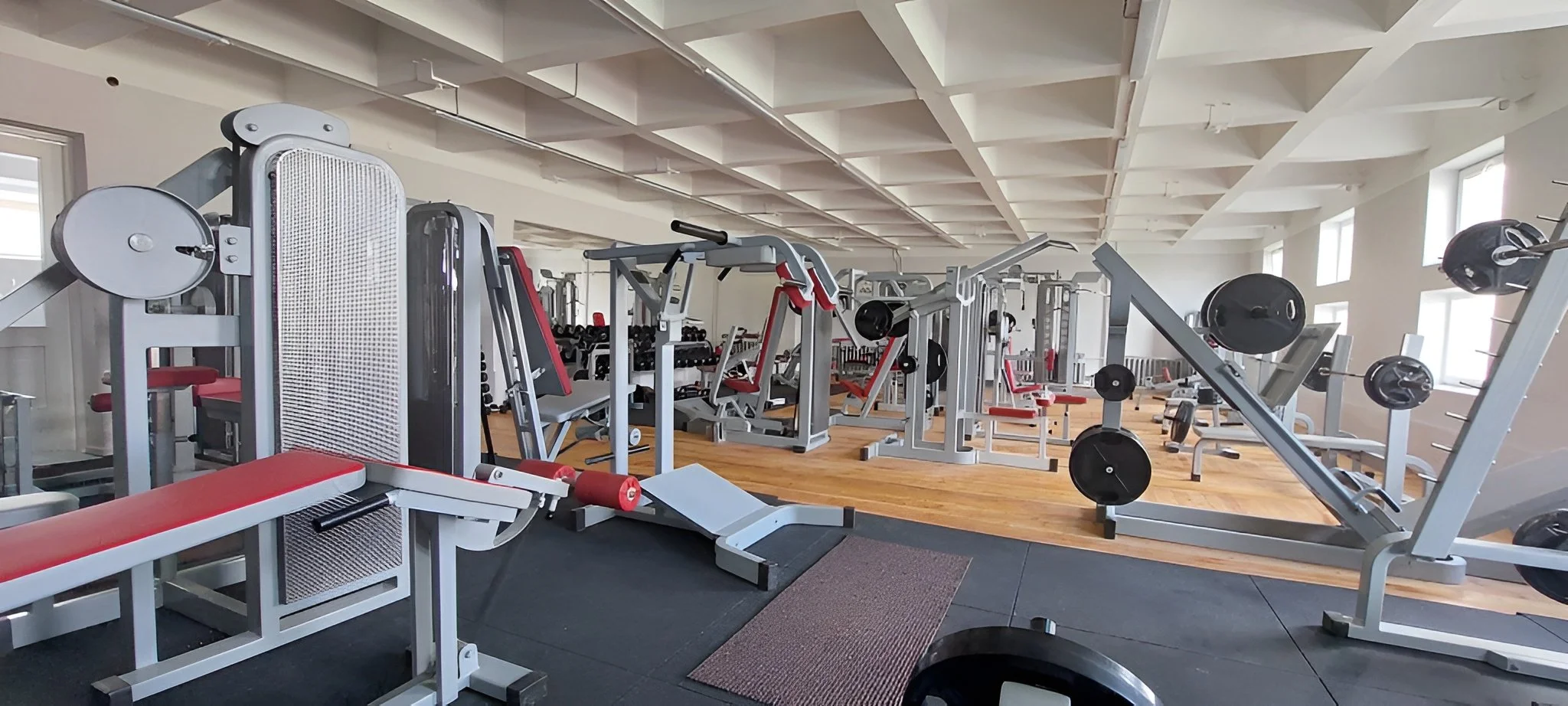Treniruoklių salė su svorių mašinomis, laisvaisiais svarmenimis ir suolais, dideliais langais ir natūralia šviesa