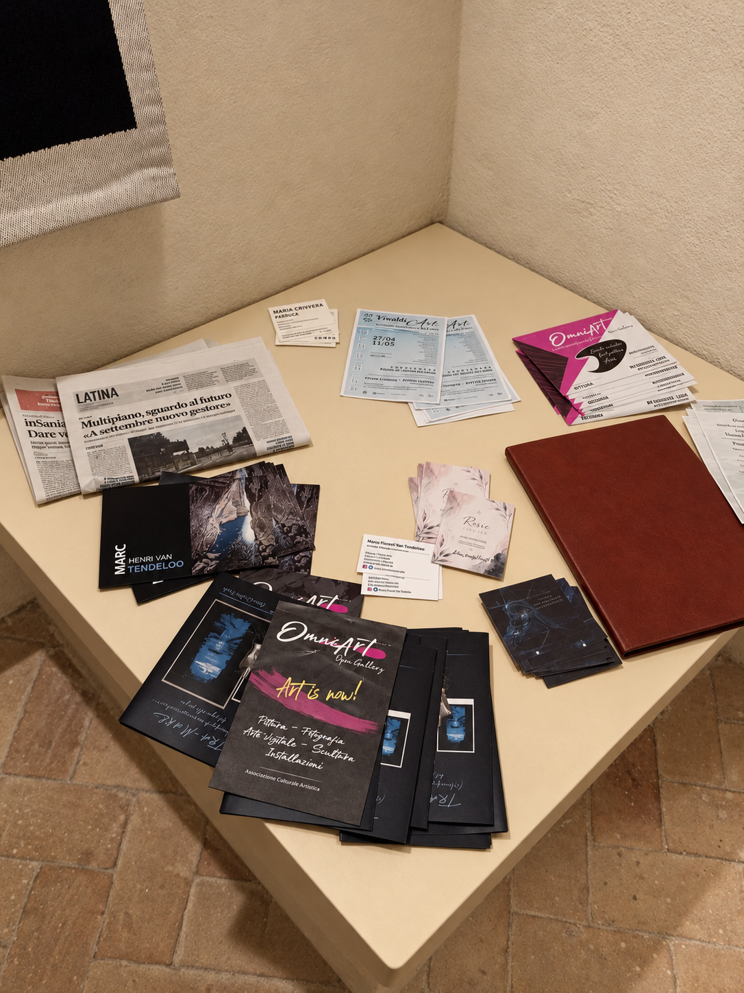 Tavolo con volantini, brochure, fogli, biglietti, e un quaderno, distribuiti su una superficie chiara, vicino a un muro beige e una tessera plastificata.