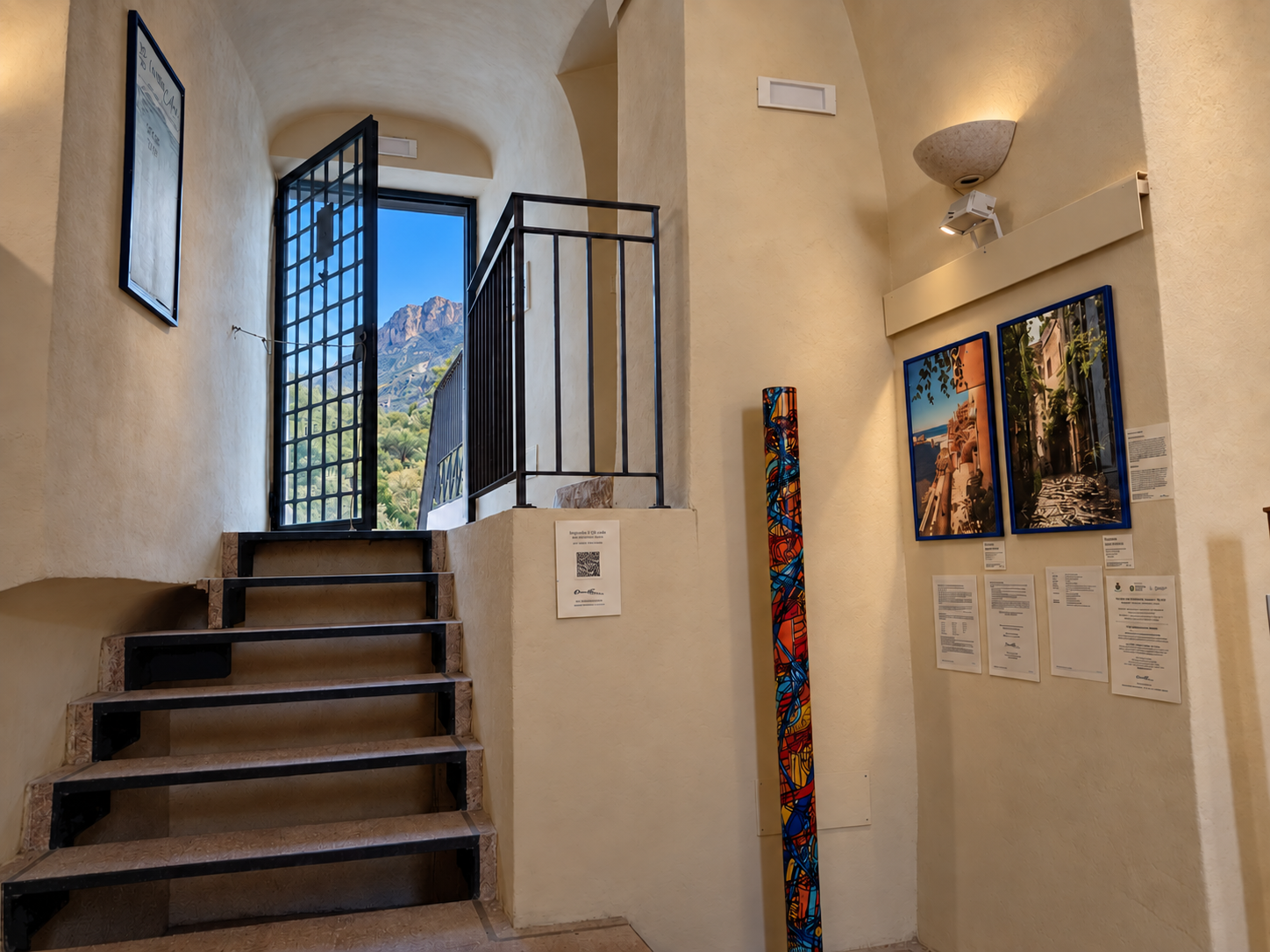 Interno di una galleria d'arte con pareti beige, dipinti e fotografie esposte, una scala in marmo con bordo in metallo nero, una finestra con sbarra che mostra montagne e natura.