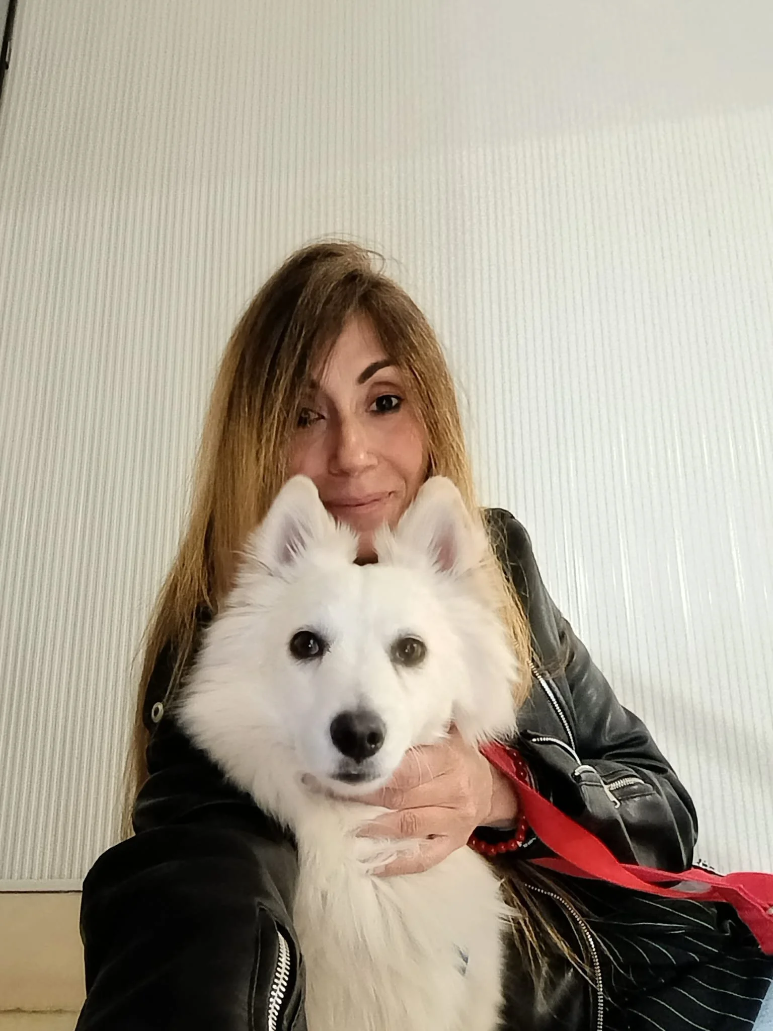 Una donna con capelli castani lunghi sorride mentre tiene un cane di razza Husky bianco con occhi diversi, uno blu e uno marrone, davanti a uno sfondo beige.