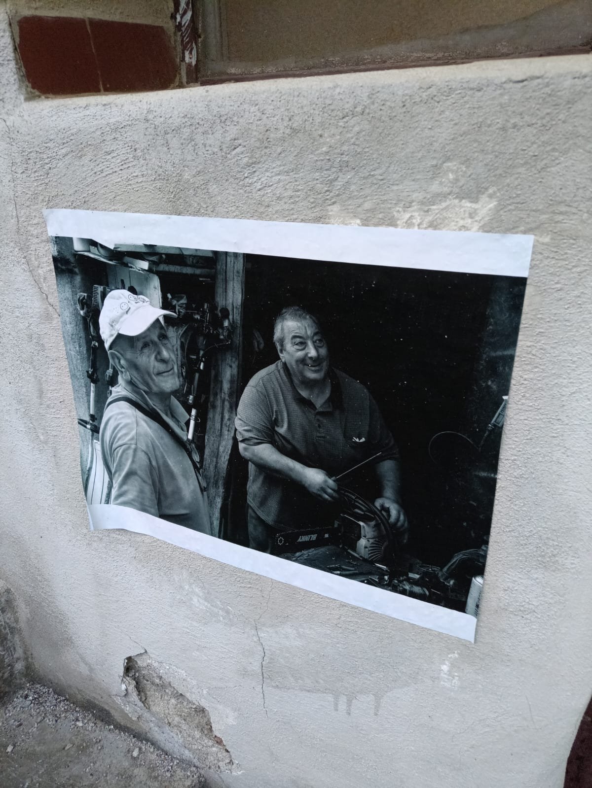 Fotografia in bianco e nero di due uomini anziani sorridenti, uno indossa un cappello e l'altro tiene una motosega, sul muro di una casa.