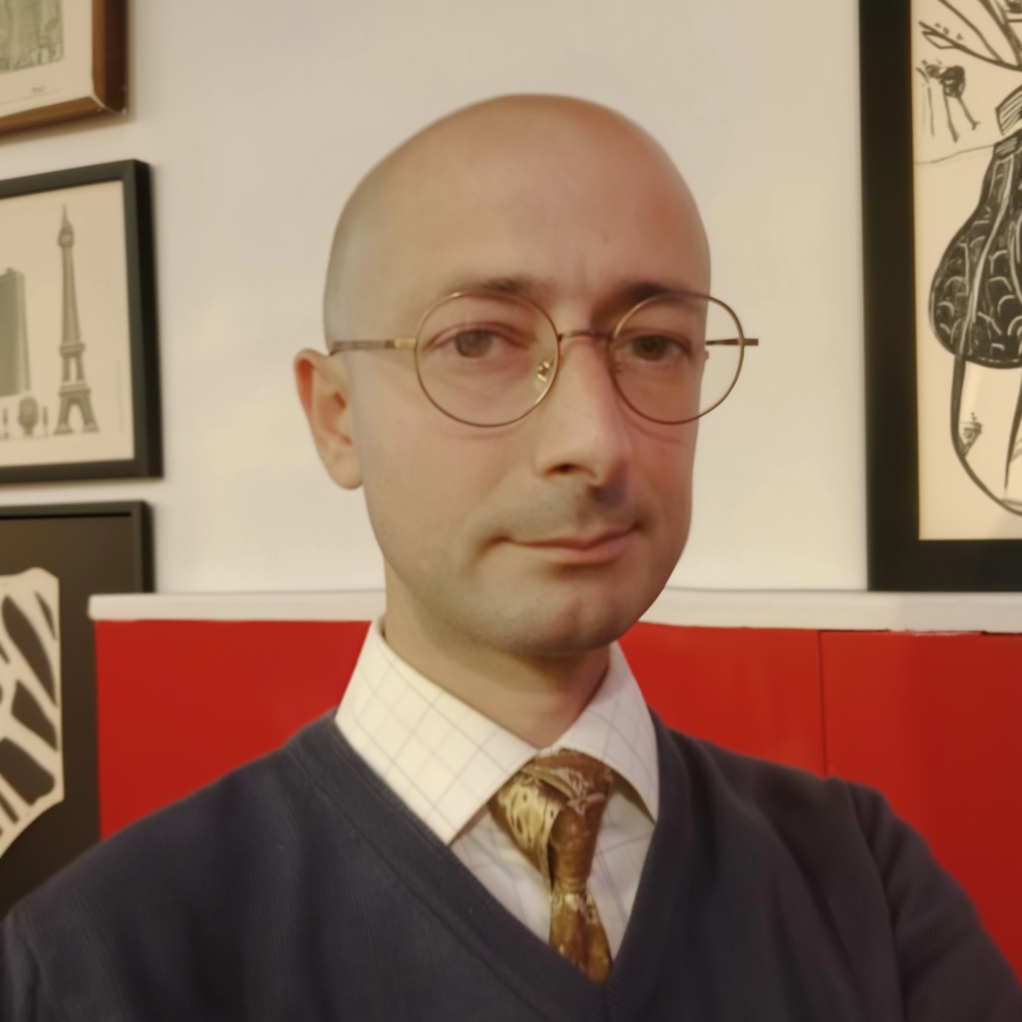 Ritaglio di un ritratto di un uomo calvo che indossa occhiali rotondi, con sfondo di pareti decorate con quadri e dipinti.