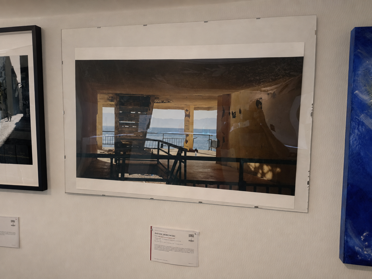 Fotografia di un ambiente interno con una vista sul mare attraverso un'apertura. La scena include strutture in legno e una ringhiera, con un paesaggio marino in lontananza.
