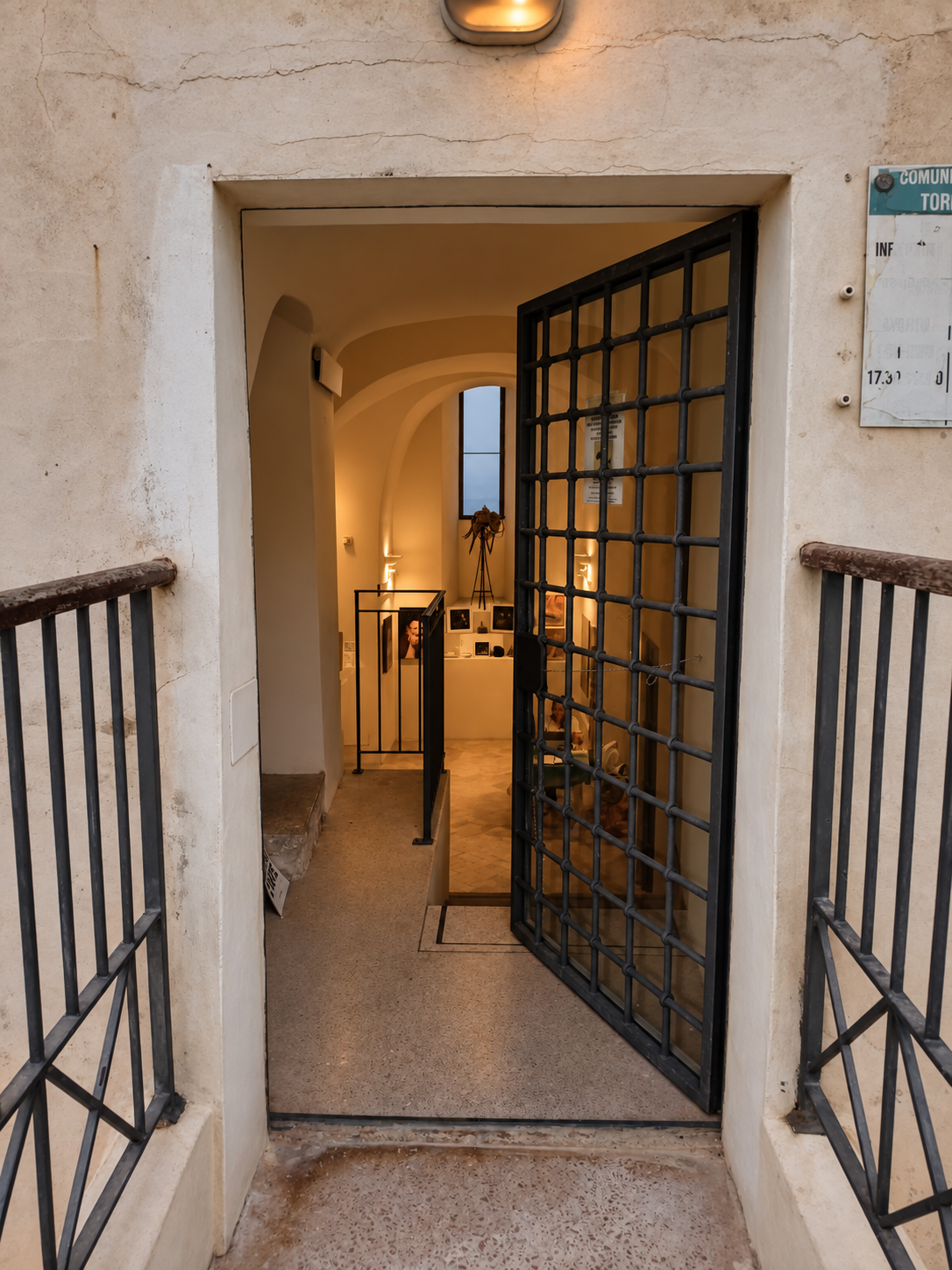 Veduta dell'ingresso di un edificio con una porta aperta di grate di ferro, che conduce a un interno con pareti chiare, decorato con luci calde, fotografie e oggetti su scaffali e tavoli.