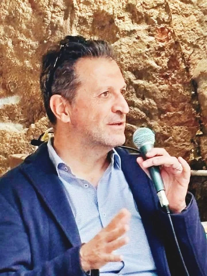 Uomo che parla in pubblico con microfono, sfondo roccioso.