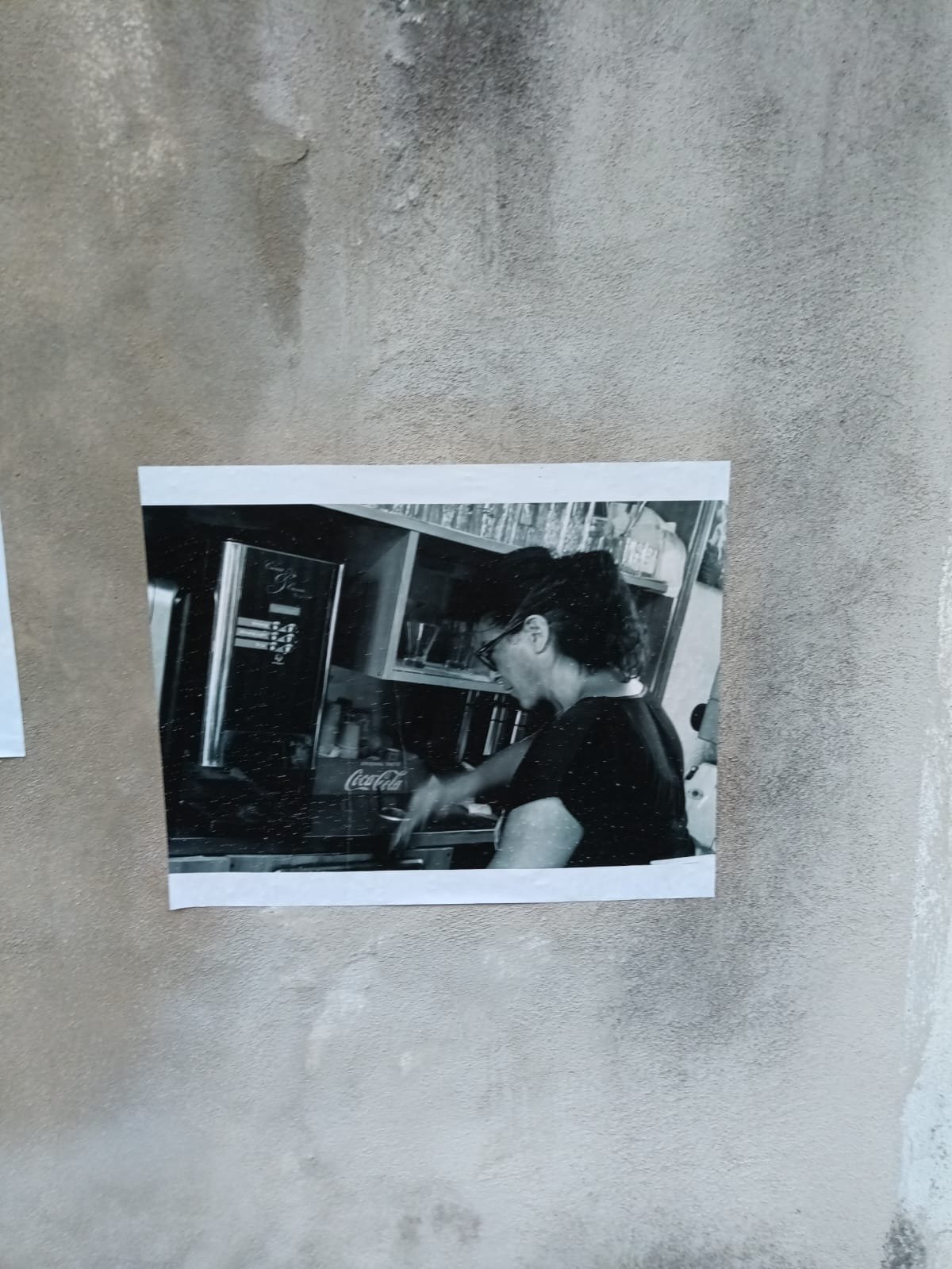 Foto in bianco e nero di una donna che prepara una bevanda dietro il bancone di un bar o caffetteria.