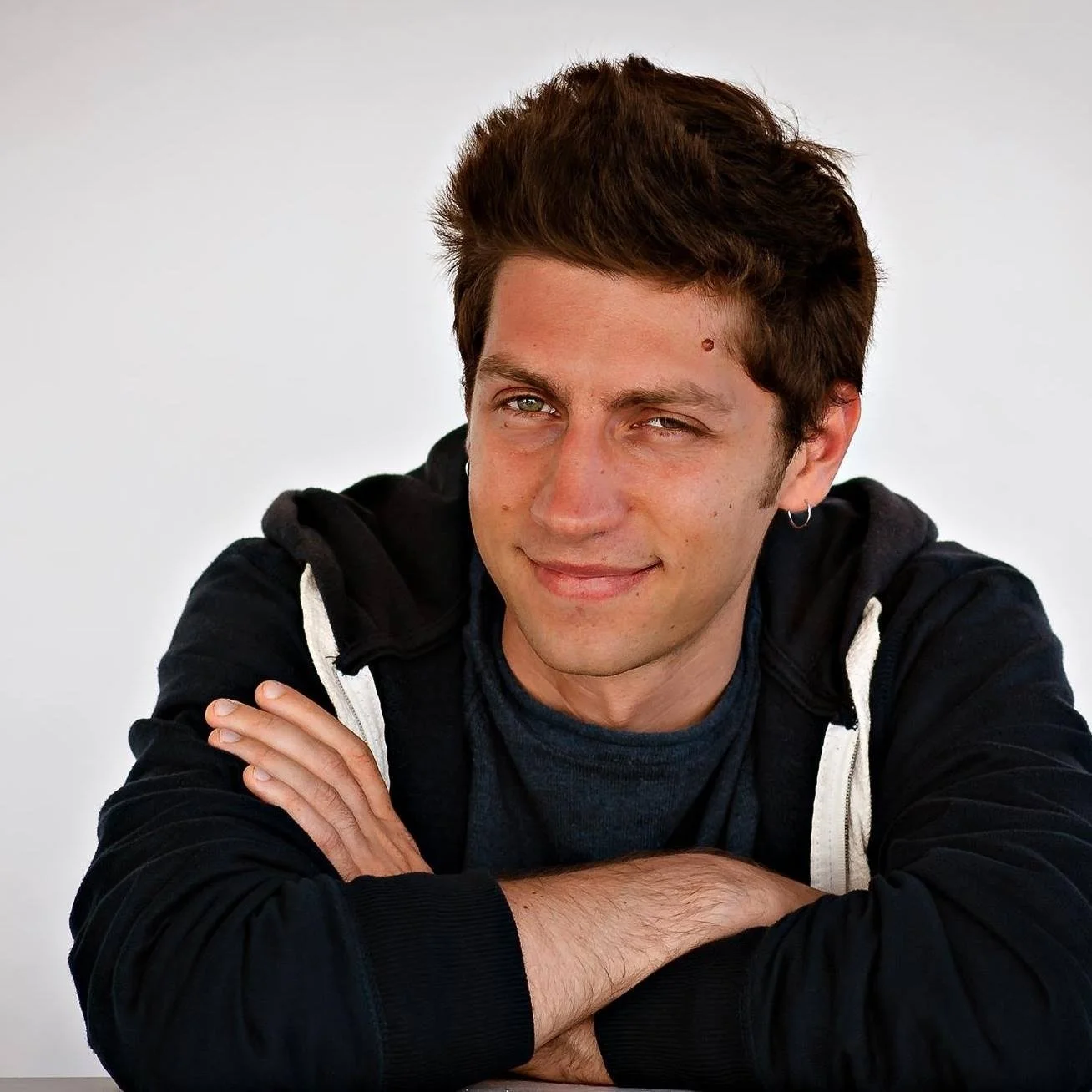 Ragazzo con capelli castani e occhi chiari sorride, indossa felpa nera con cappuccio e braccia incrociate, sfondo bianco