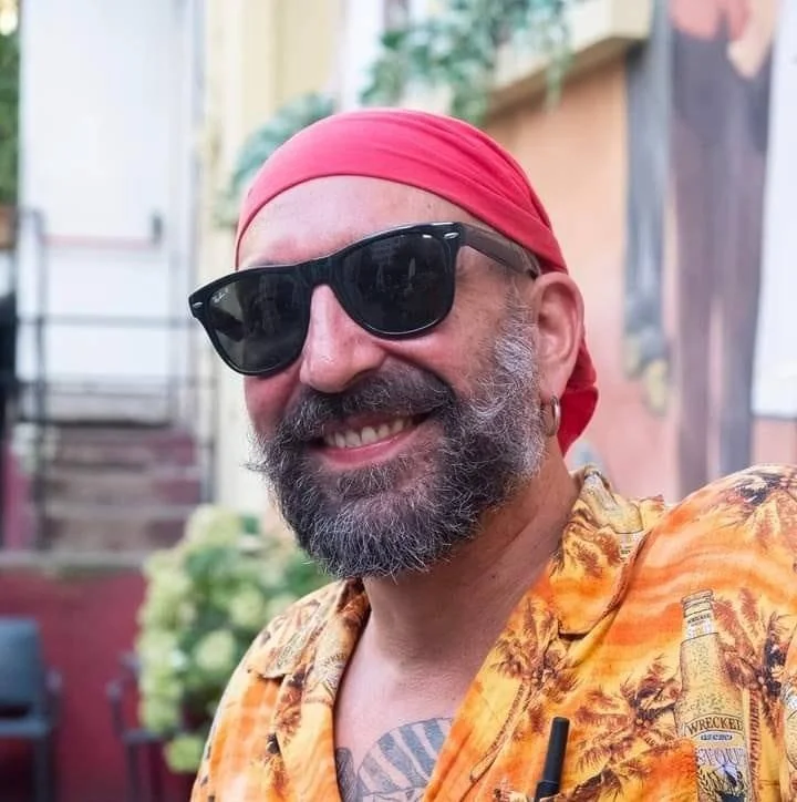 Uomo sorridente con barba e baffi, indossa occhiali da sole neri e una bandana rossa, vestito con una camicia hawaiana arancione con motivi tropicali, sullo sfondo un ambiente esterno con piante.