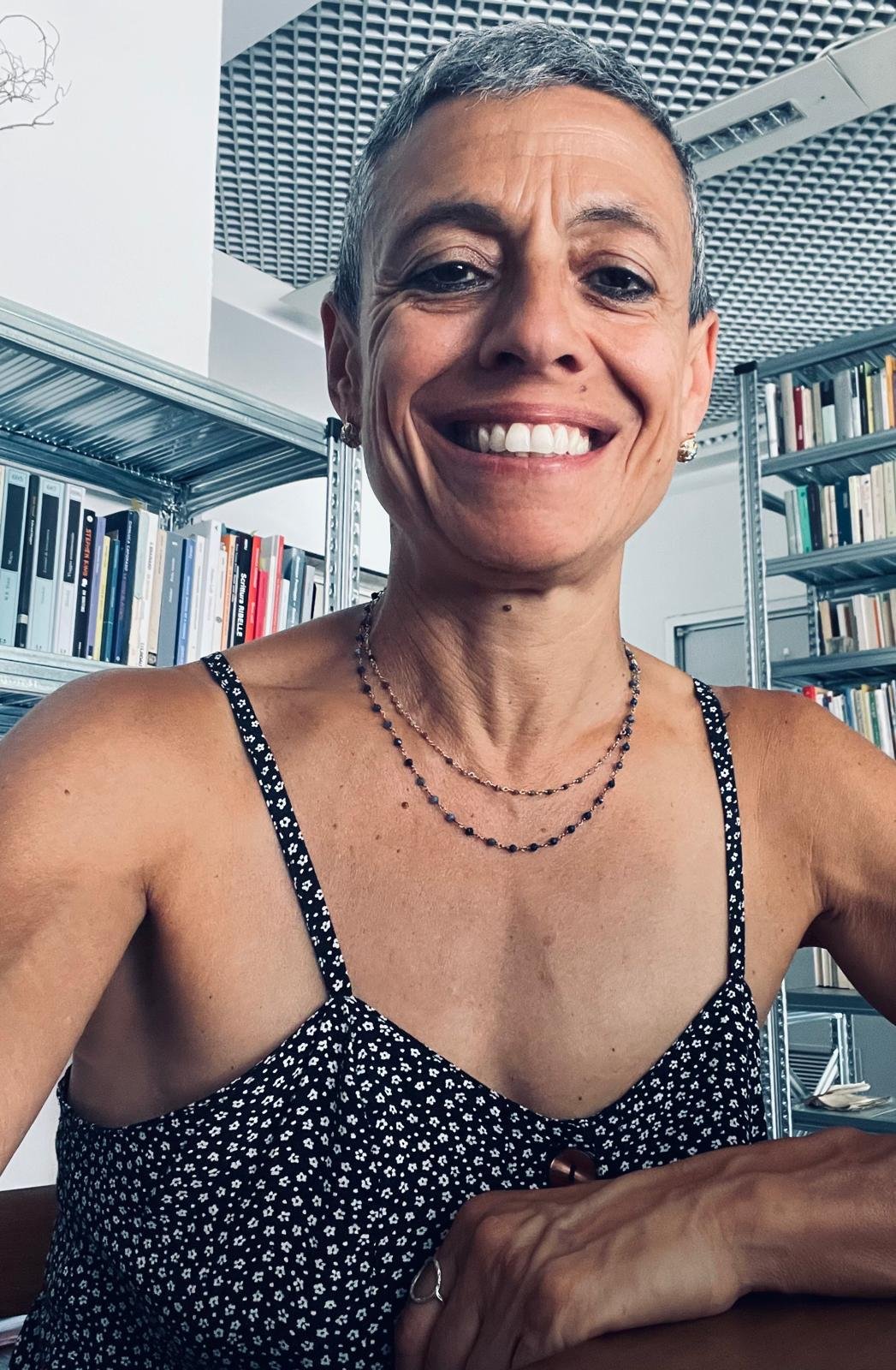 Una donna sorridente in un ambiente con scaffali pieni di libri, indossa un vestito nero con pois bianchi e collane. È all'aperto o in un luogo moderno, con un soffitto a griglia.