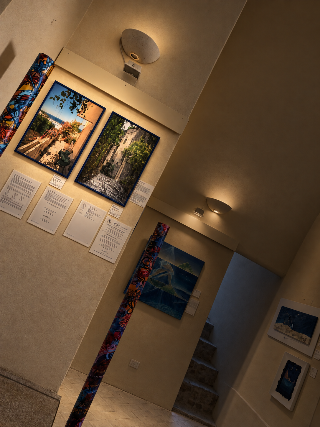 Mostra d'arte in una galleria con dipinti e fotografie appesi alle pareti, illuminata da luci da soffitto
