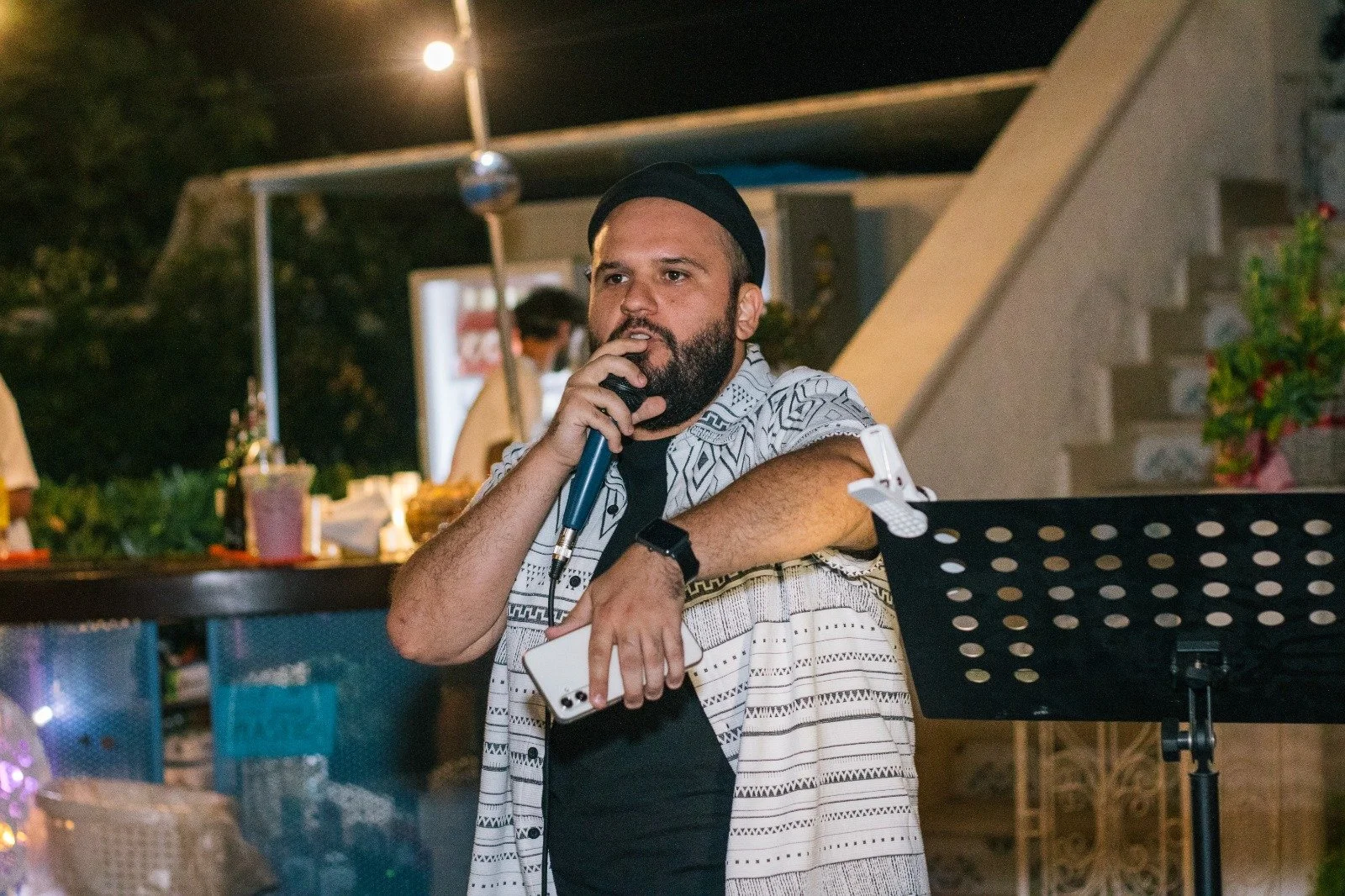 Uomo con barba che parla in microfono in un evento notturno, con sfondo di bar e luci.