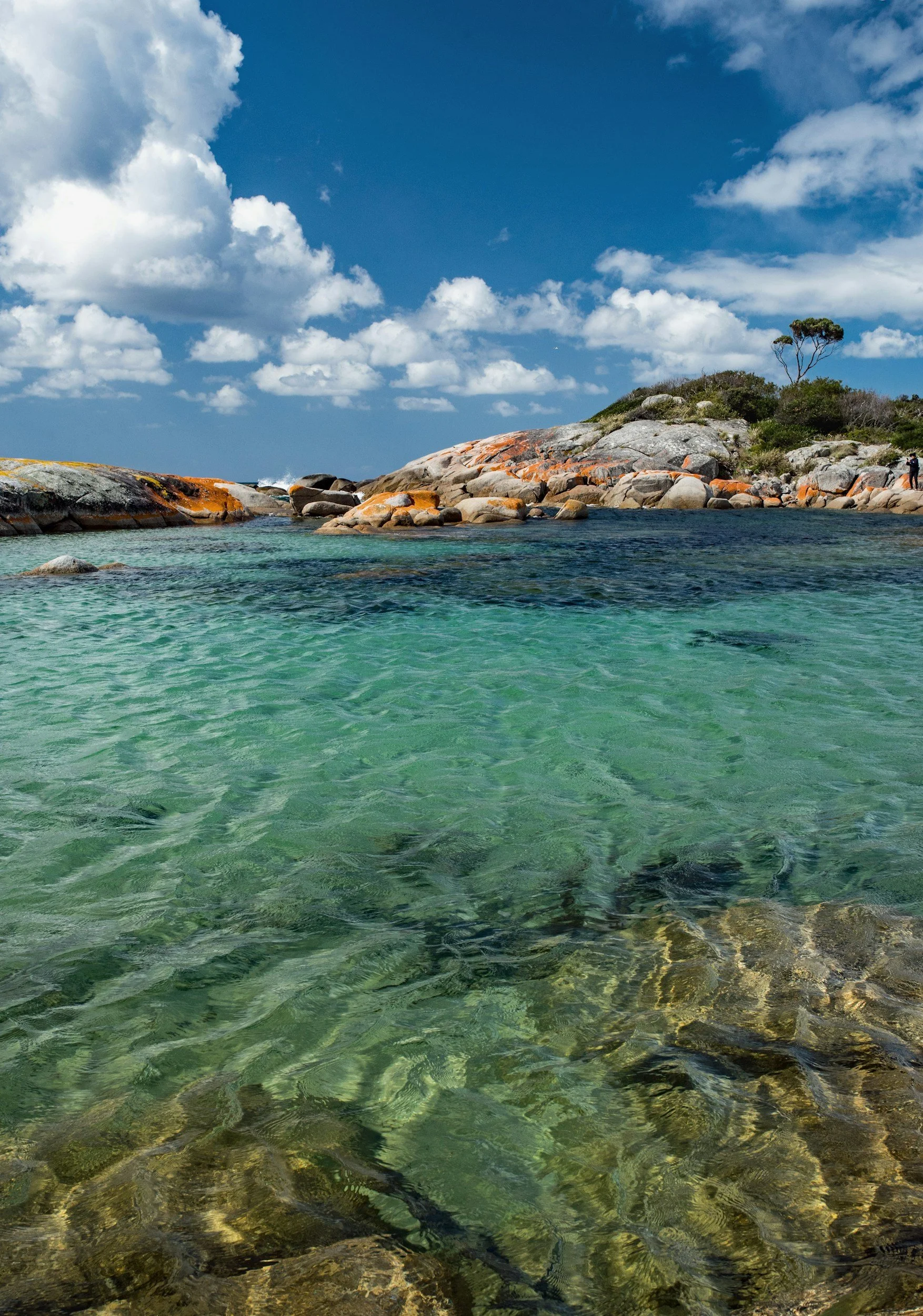 Clear turquoise water with rocks and a rocky coastline under a partly cloudy blue sky.