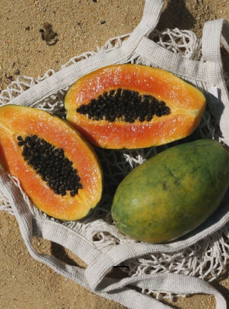 Two halves of a papaya with black seeds and one whole green papaya on a netted bag on sandy ground.