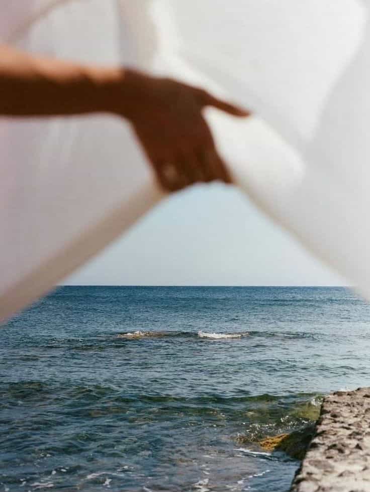 A person’s hand and arm reaching through or around a white structure, with a view of the ocean and a rocky shoreline in the background.