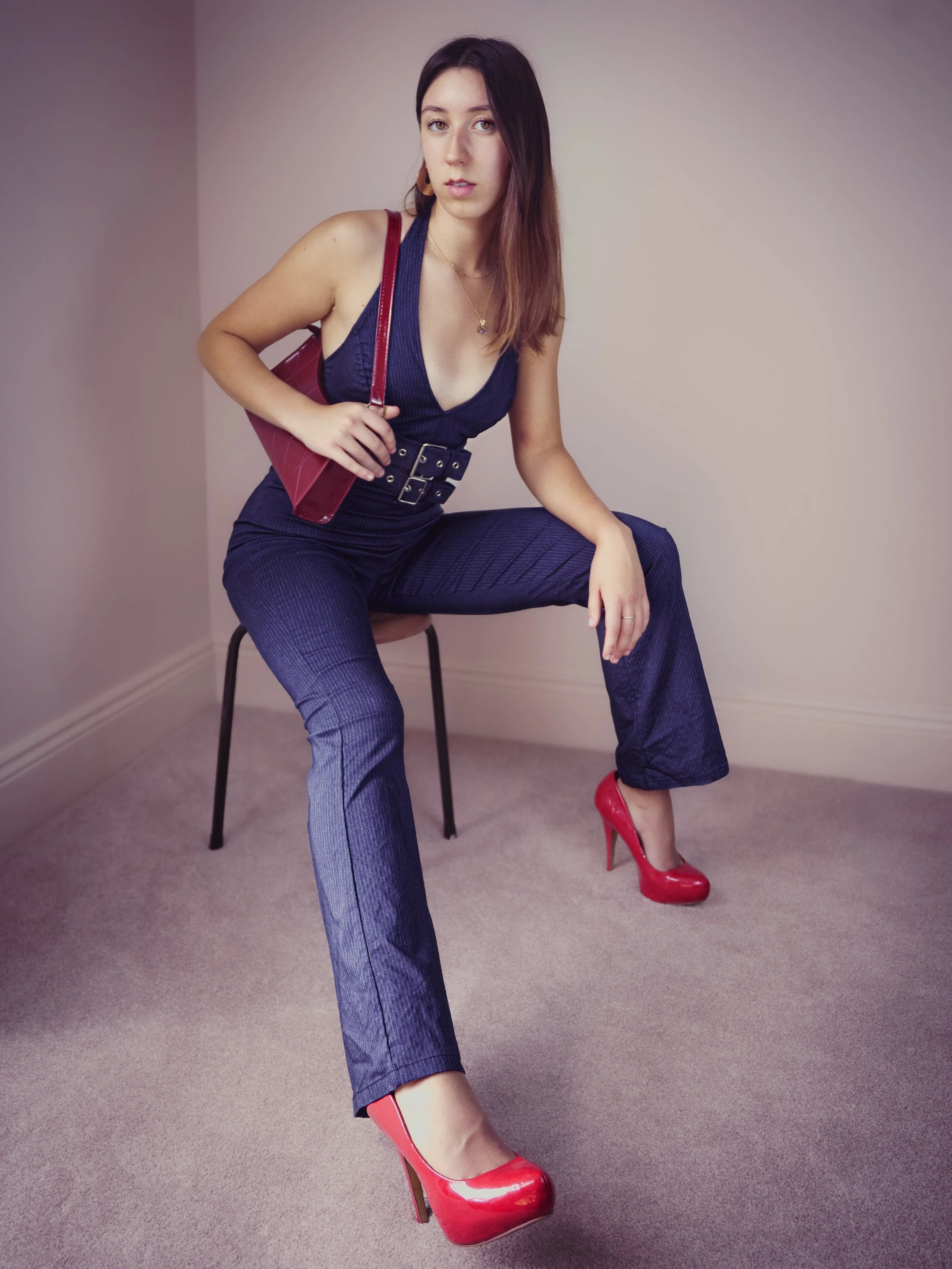 Woman sitting on a chair wearing a dark blue jumpsuit, red high heels, and carrying a red purse, with neutral expression and long brown hair.