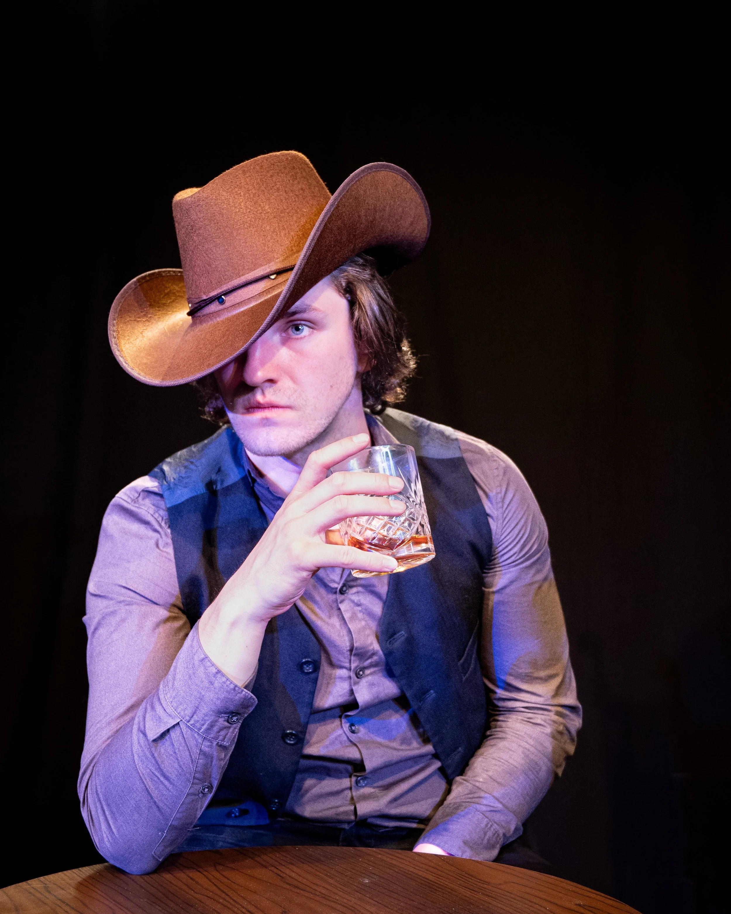 A man wearing a cowboy hat and a brown shirt, sitting at a wooden table, holding a glass of whiskey, in a dark setting.