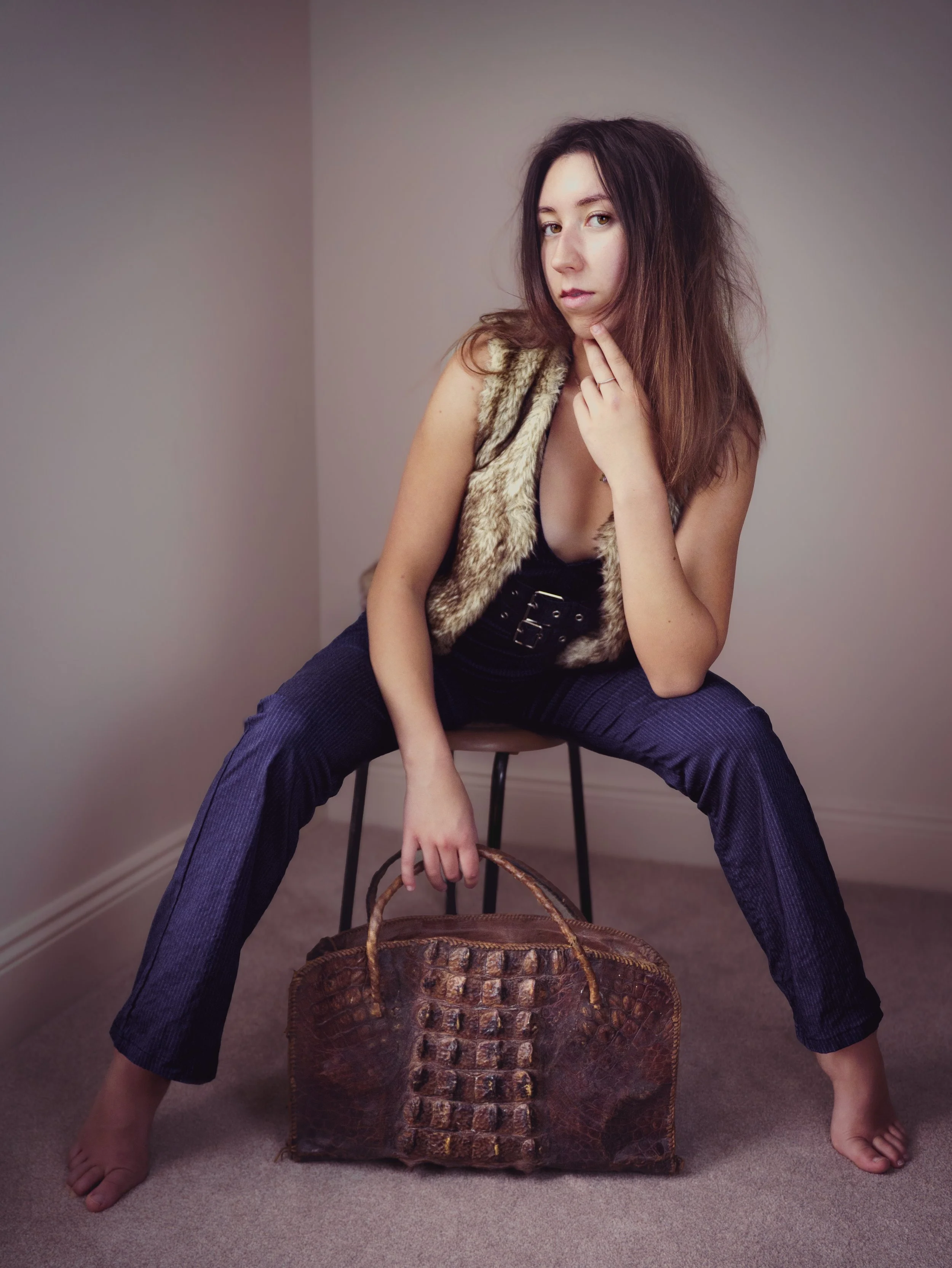 A young woman with long, tousled hair sitting on a chair in a corner of a room. She is wearing dark pants, a fur vest, and a black top, and is holding a large brown crocodile-embossed handbag. She is barefoot and has a contemplative expression, with