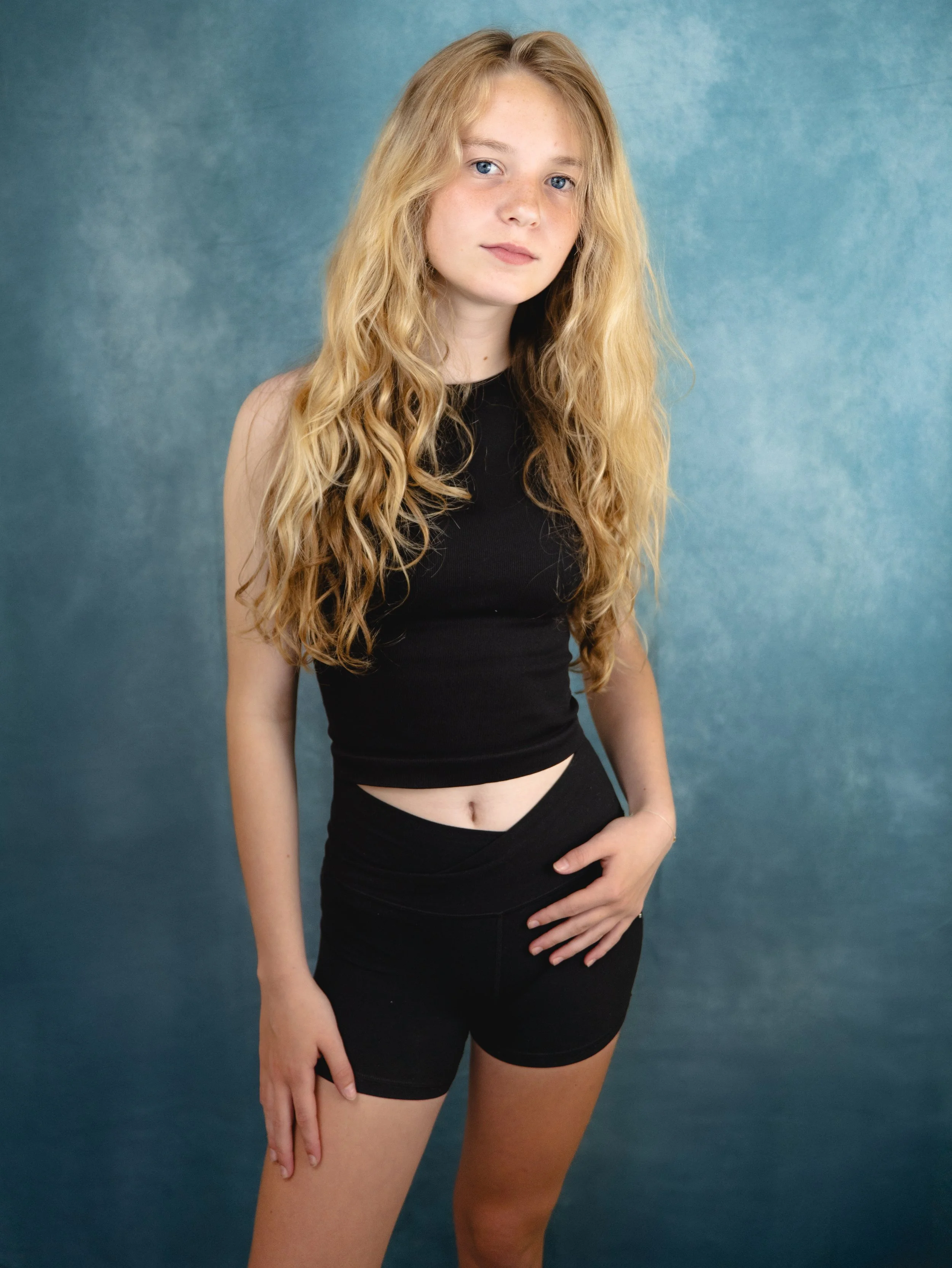 Young woman with long wavy blonde hair and light skin, wearing a black sleeveless crop top and black shorts, standing against a textured blue background.