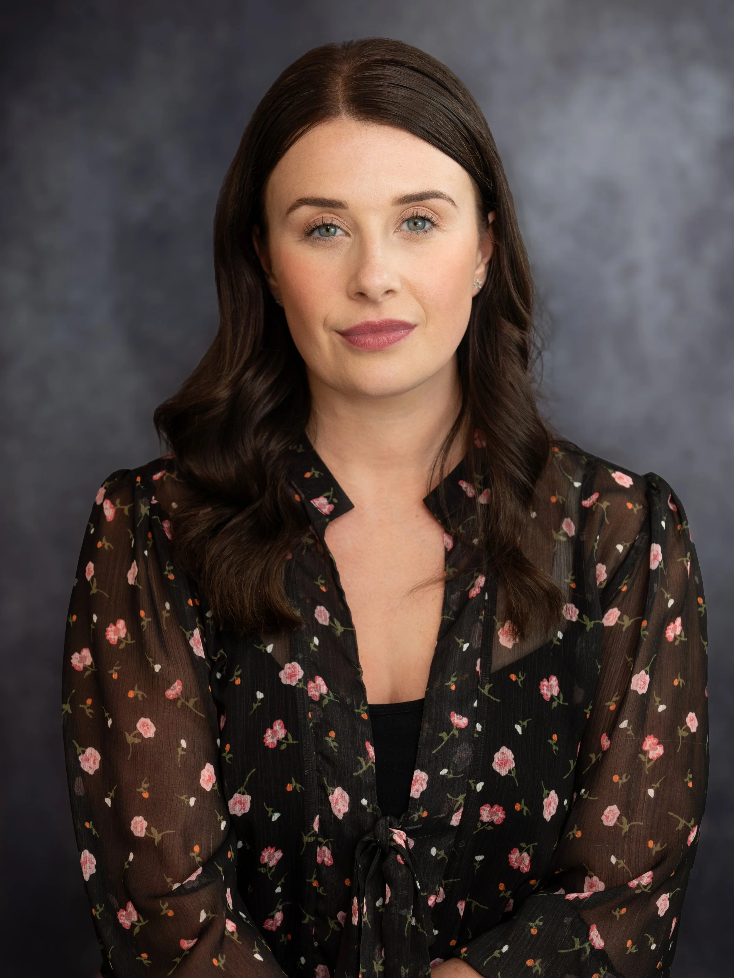 An actors headshot of a woman in a black blouse, with long dark brown hair. She sits in front of a dark grey background with a steely expression. 