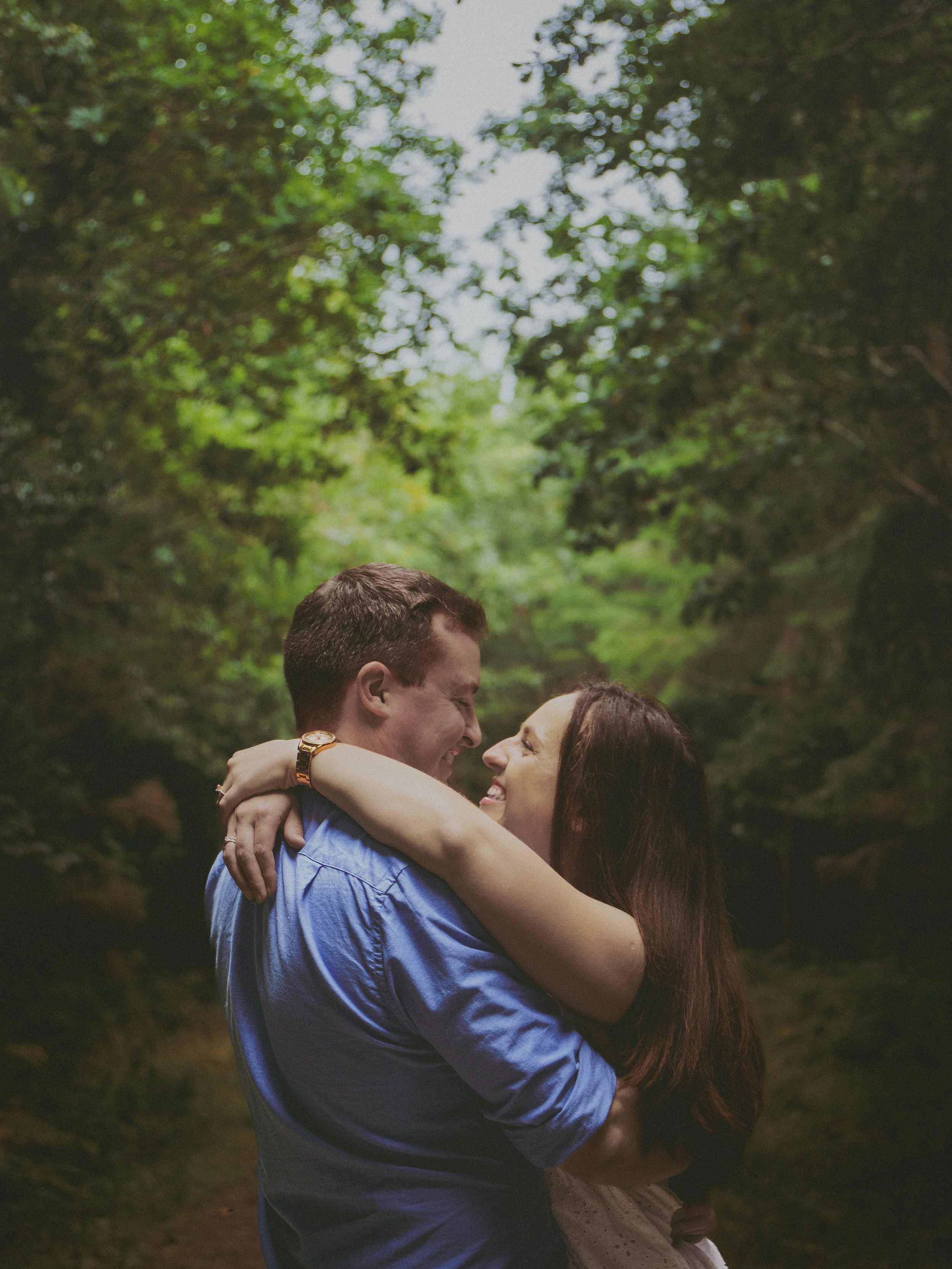 A couple embracing outdoors in a green, wooded area, smiling and facing each other closely.