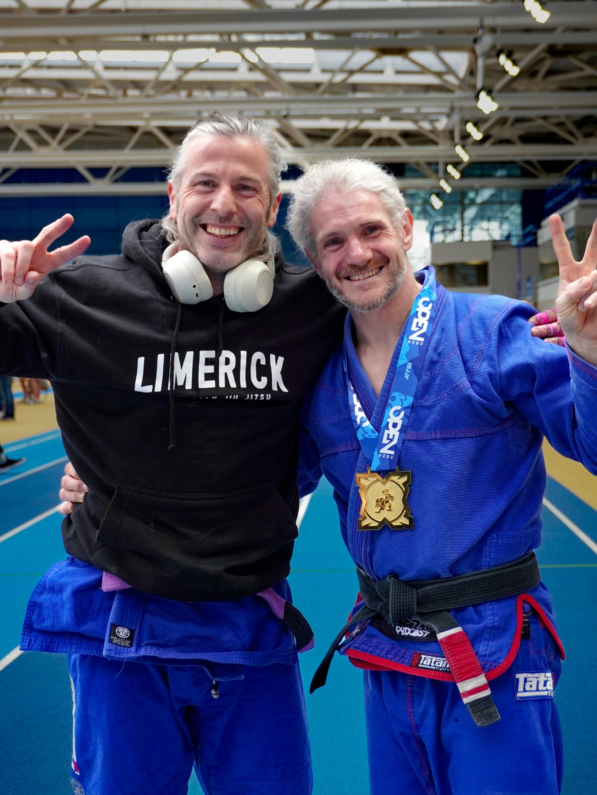 Two men smiling and making victory signs with their hands at a sports event, one wearing a black Limerick hoodie and the other wearing a blue gi with a medal around his neck, in a large indoor sports facility.