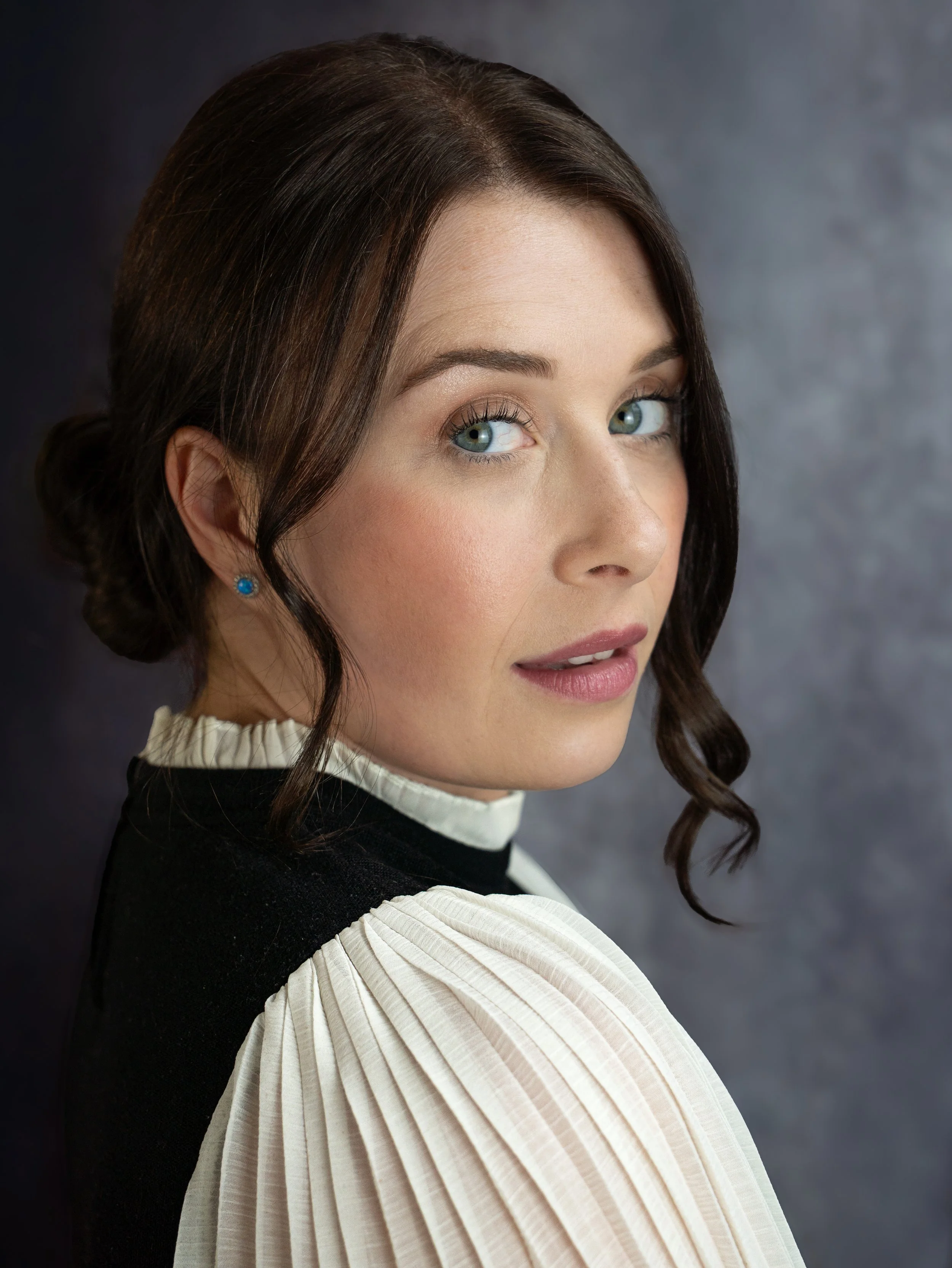 An actor headshot of a woman looking over her shoulder, she is sitting infront of a dark grey background. She has dark brown hair tied up in a bun. Her top looks like period clothing with soft cream pleated sleeves and collar. 