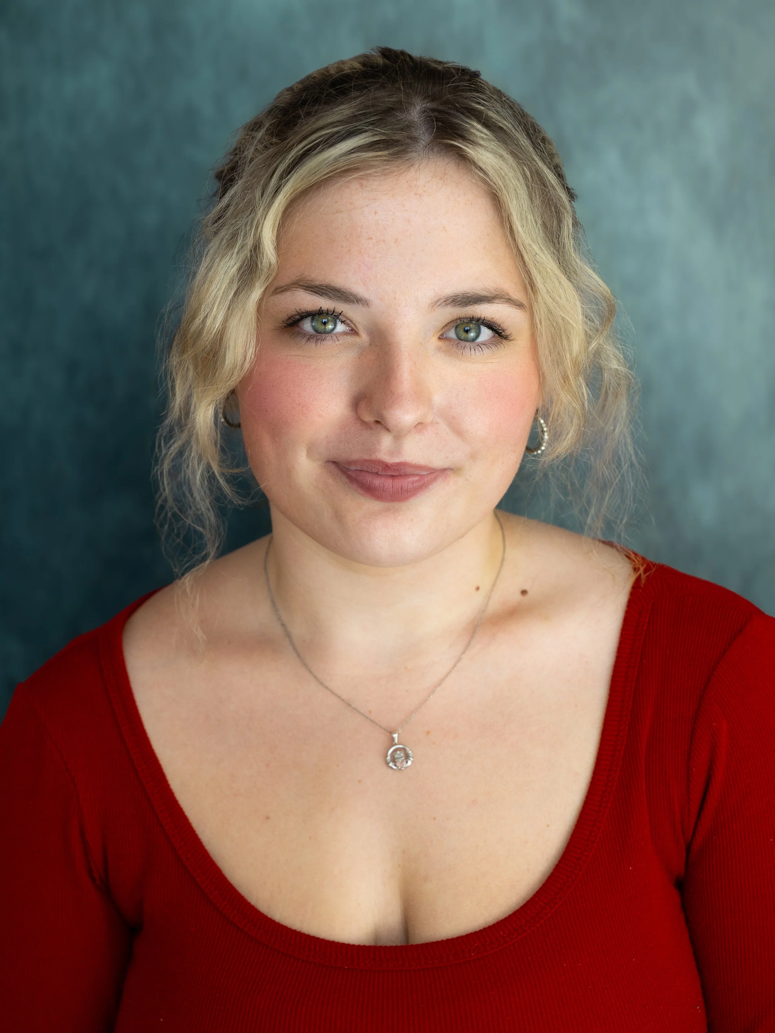 An actors headshot of a woman wearing a dark red top. Her hair is back in a ponytail with her fringe loose on either side. She has wavy blond hair, green eyes and is facing the camera with a soft smile.