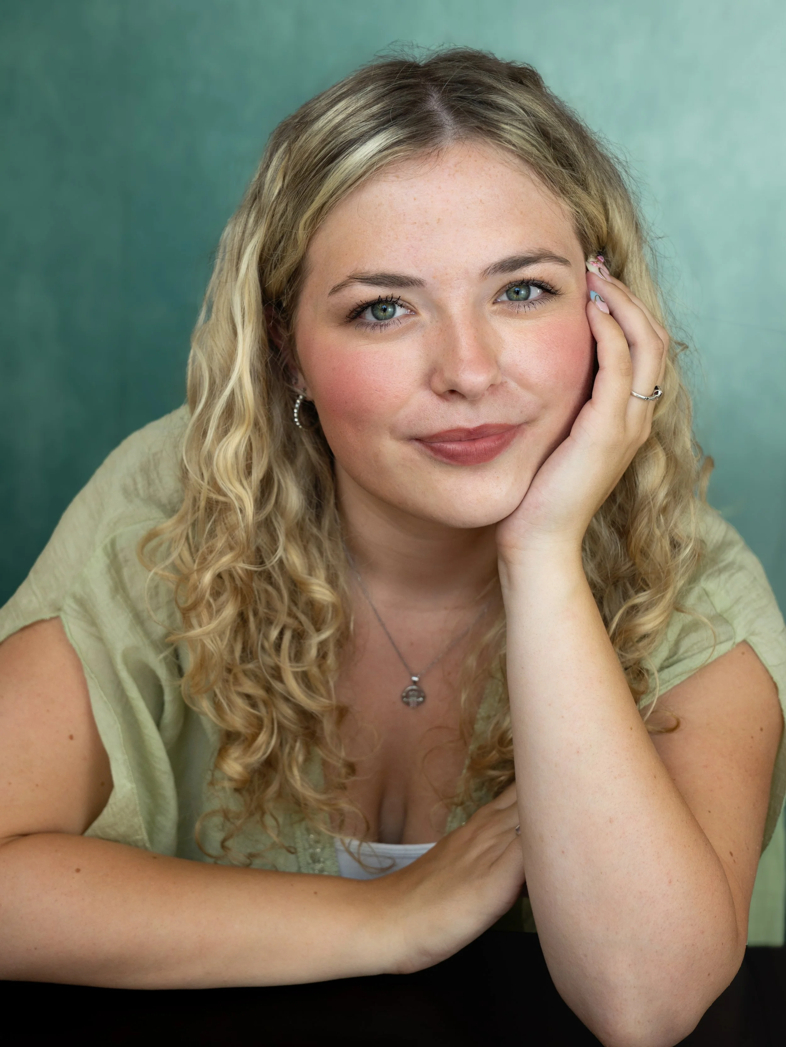 An actors heashot of a girl with blond curly hair, leaning on her left hand. She is wearing a light green top, she has green eyes and is smiling softly.