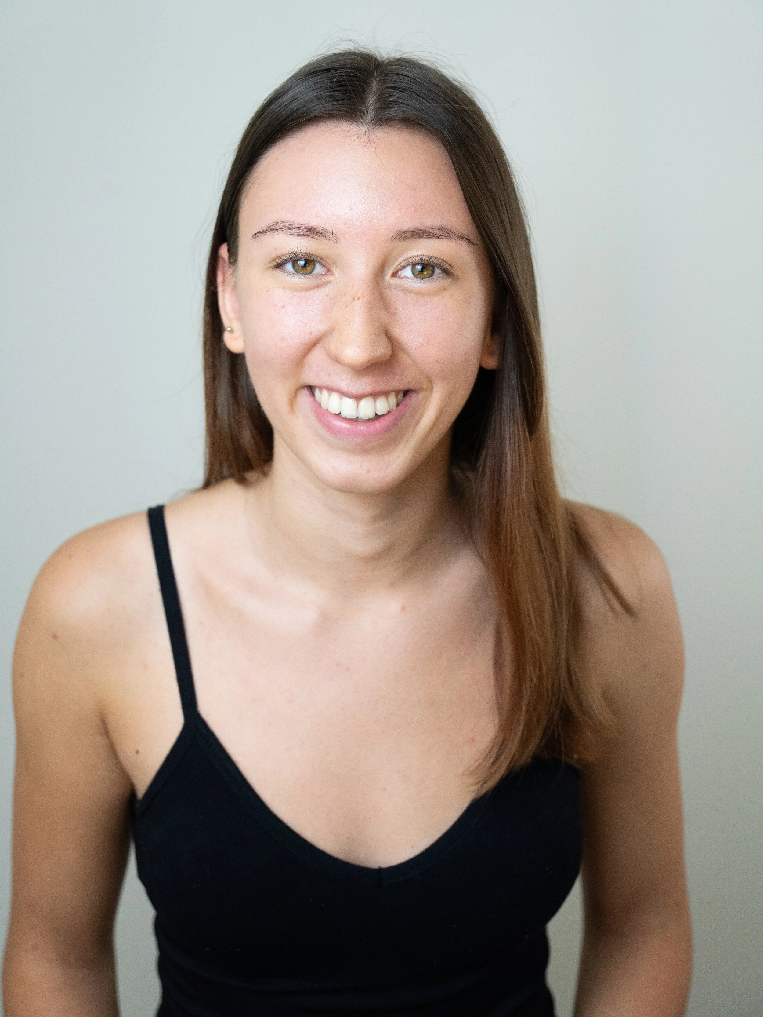 A young woman with long brown hair and light skin, smiling at the camera, wearing a black spaghetti strap top, against a plain light-colored background.