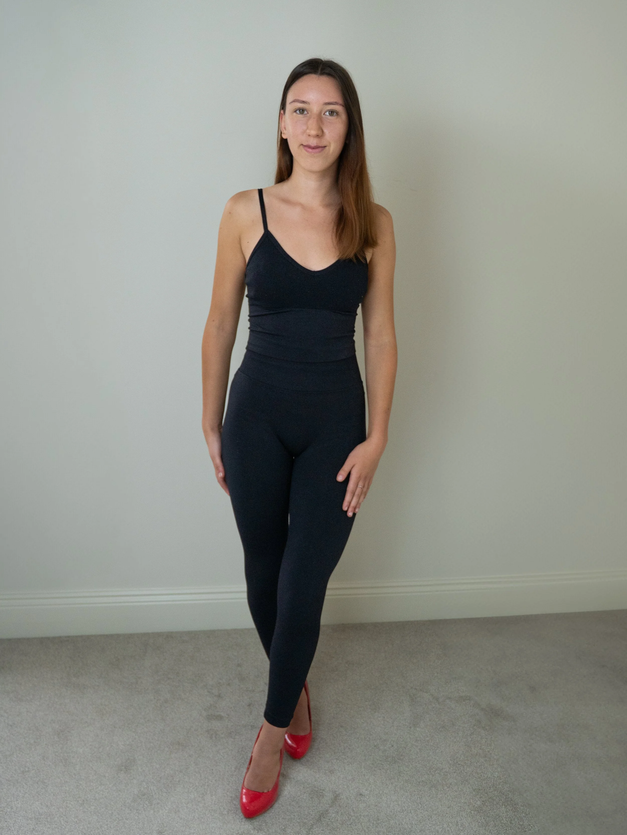 A woman standing against a plain light-colored wall, wearing a black tank top, black leggings, and red high heels.