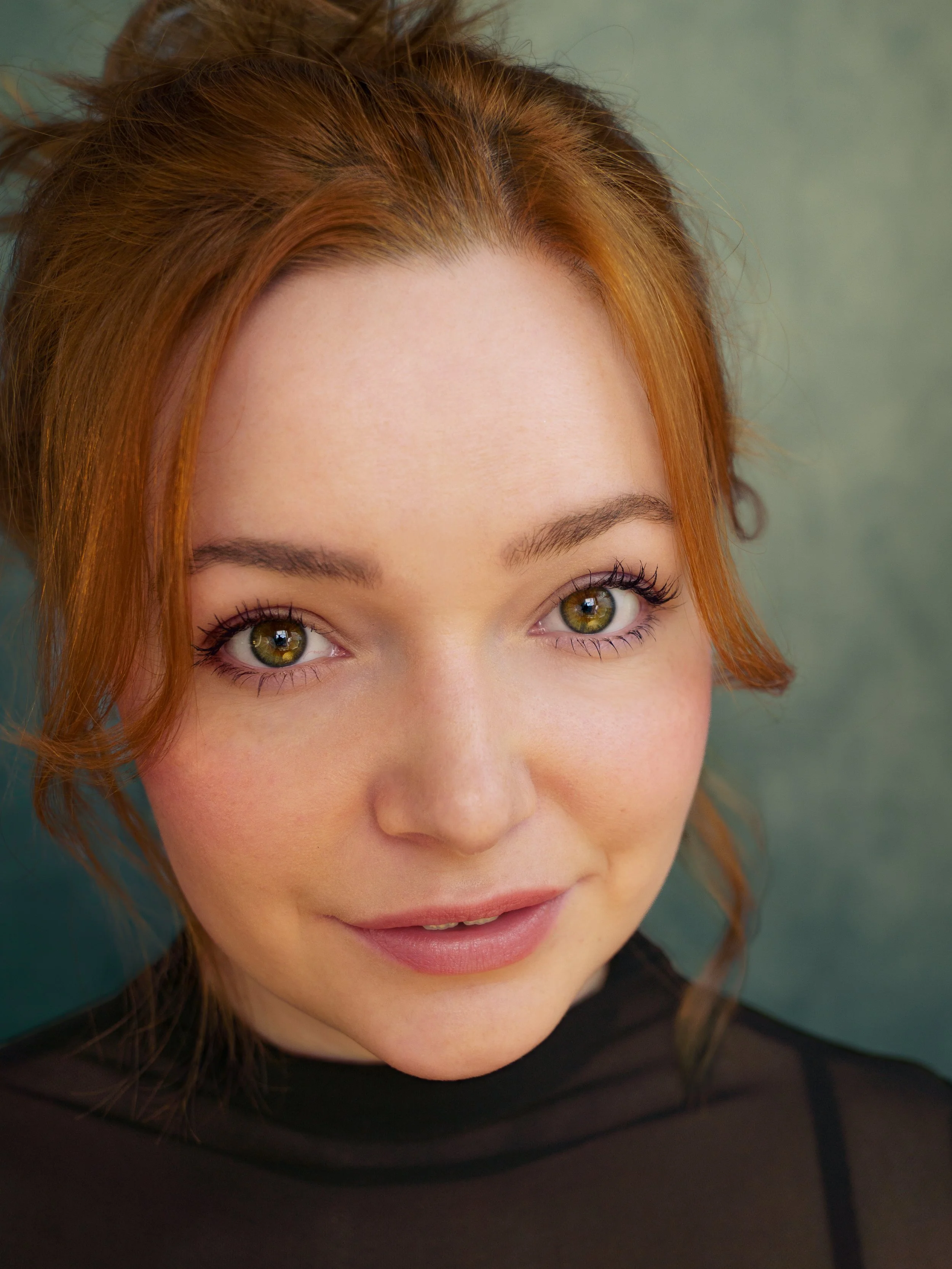 Close-up photo of a young woman with red hair, green eyes, light makeup, and a friendly expression, wearing a black top.