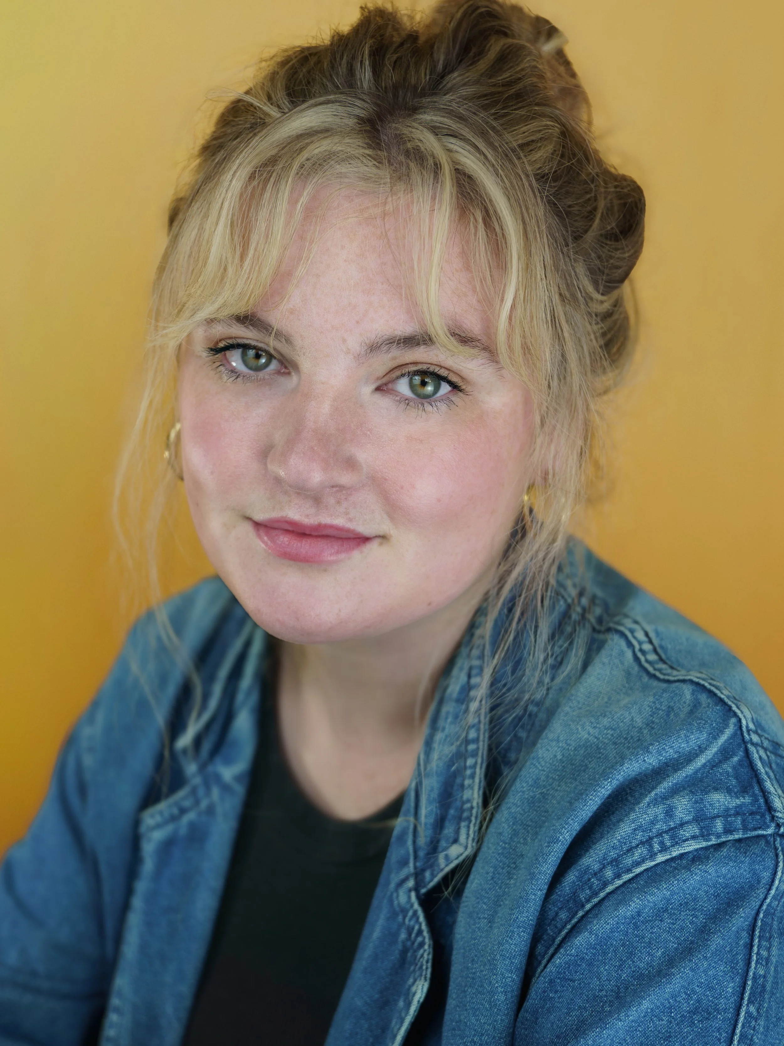 An actor headshot of a woman sitting infront of a bright yellow background. She has blond curly hair tied up in a bun with her fringe down framing her face. She is wearing a blue denim jacket and has a soft smile on her face. 