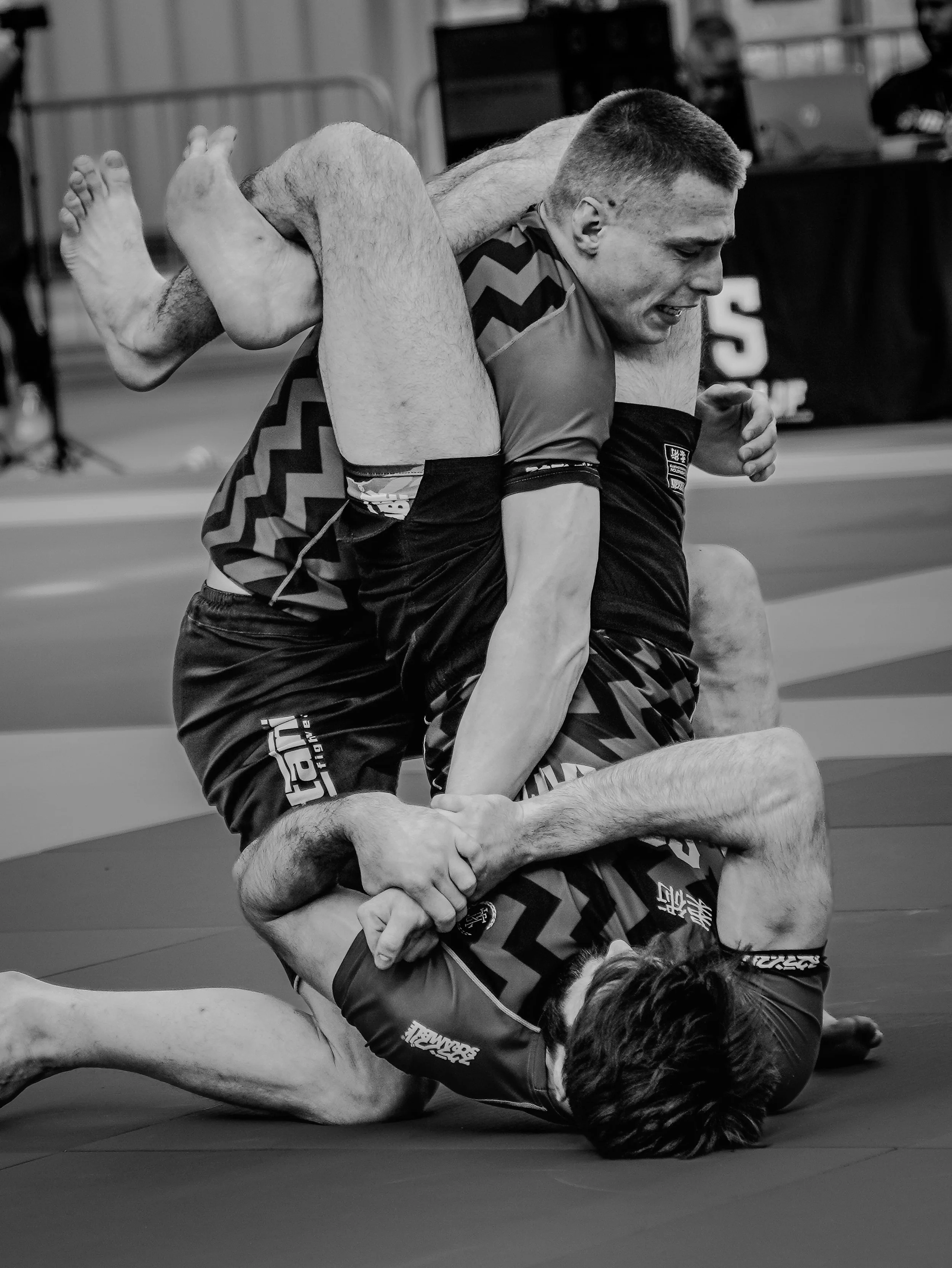 Two male mixed martial artists grappling on a mat during a match, with one on the ground and the other on top, both focused on their positions.