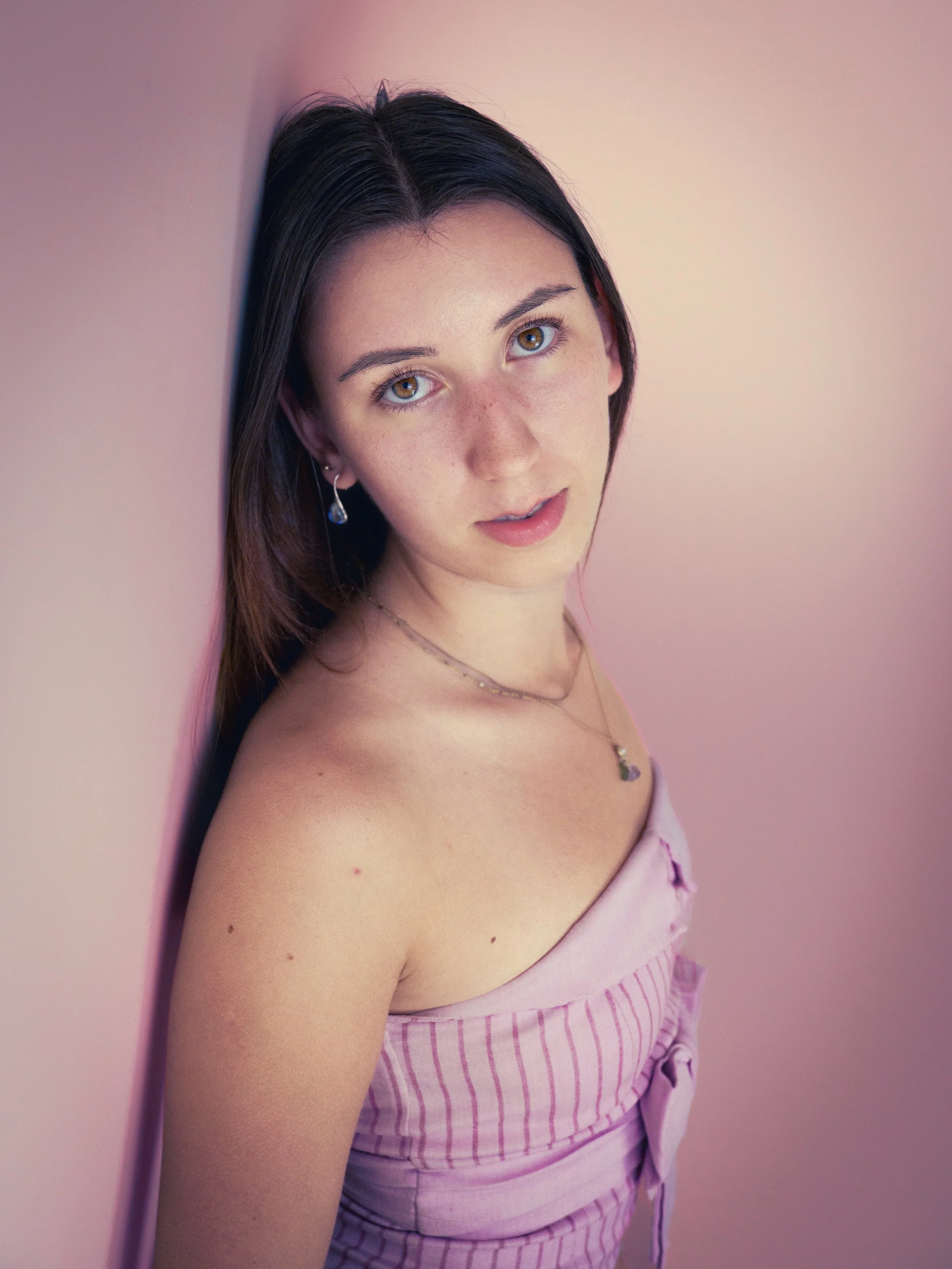 A young woman with brown hair, brown eyes, and light skin, leaning against a pink wall, wearing a pink striped top and jewelry.