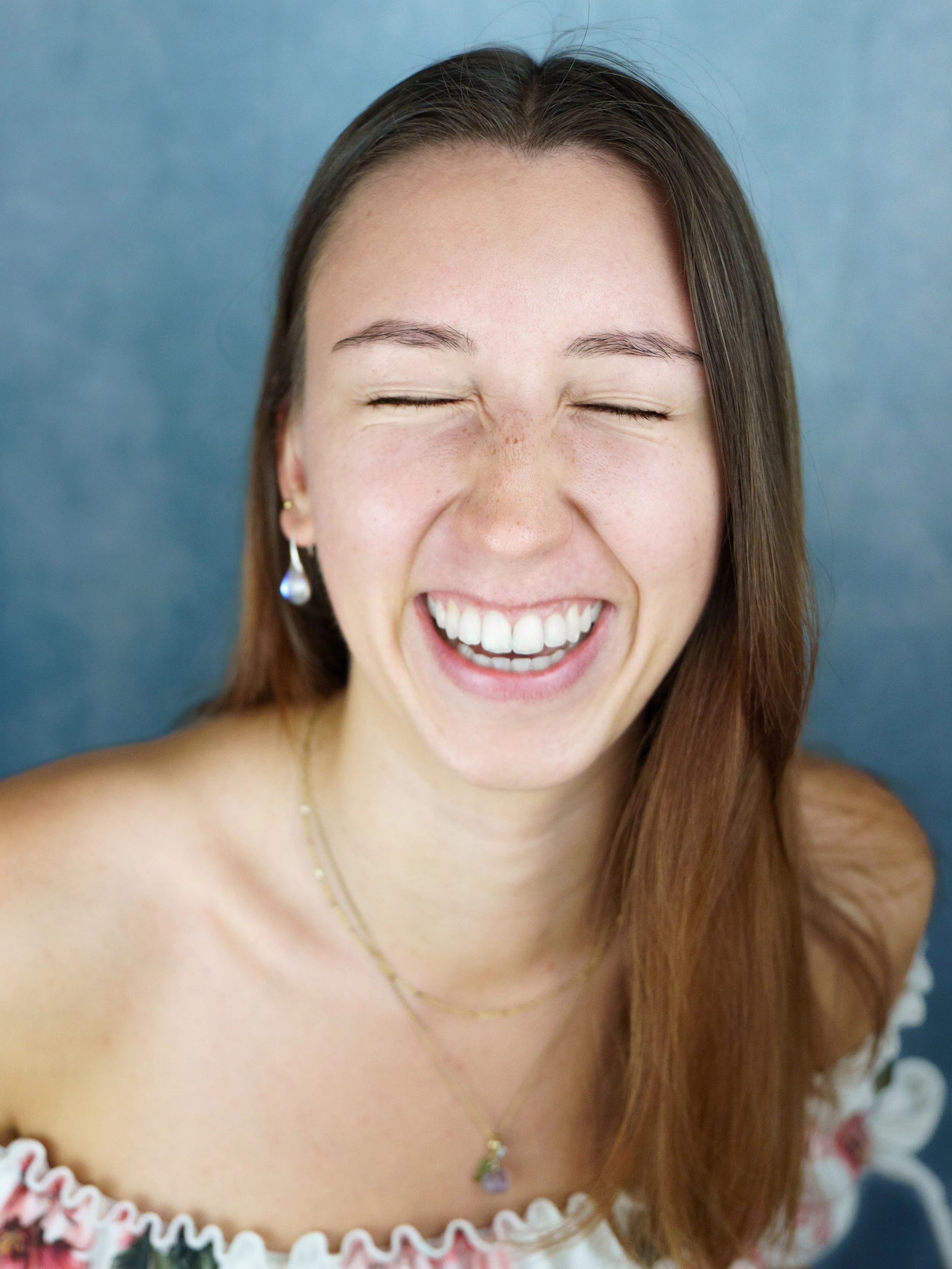 Close-up of a woman with long brown hair, smiling with eyes closed, wearing earrings, a necklace, and an off-the-shoulder top, against a blue background.