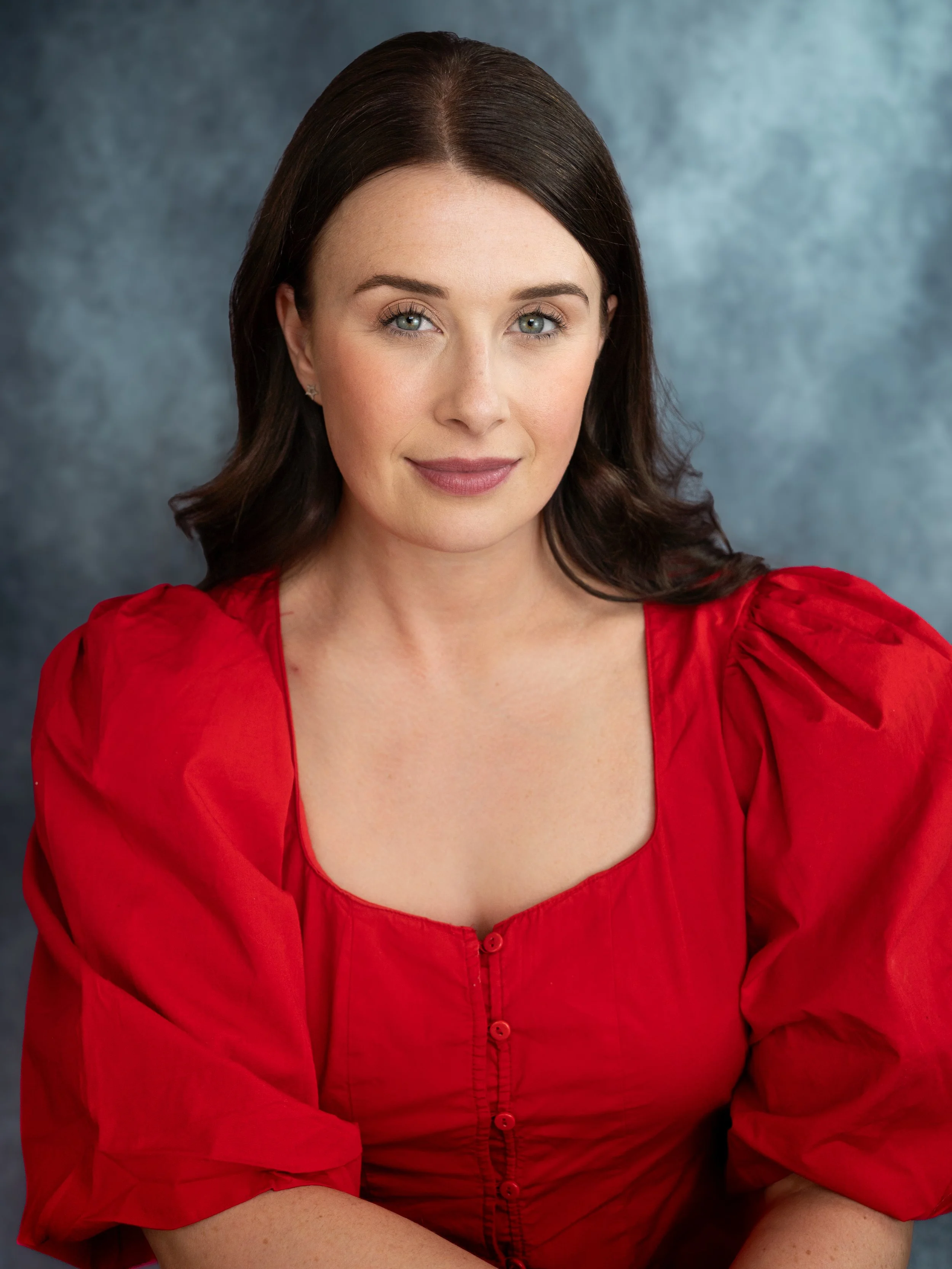 An actors headshot of a woman in a bright red top sitting infront of a blue/grey background. She has sleek dark brown hair and has a slight smirk on her face.