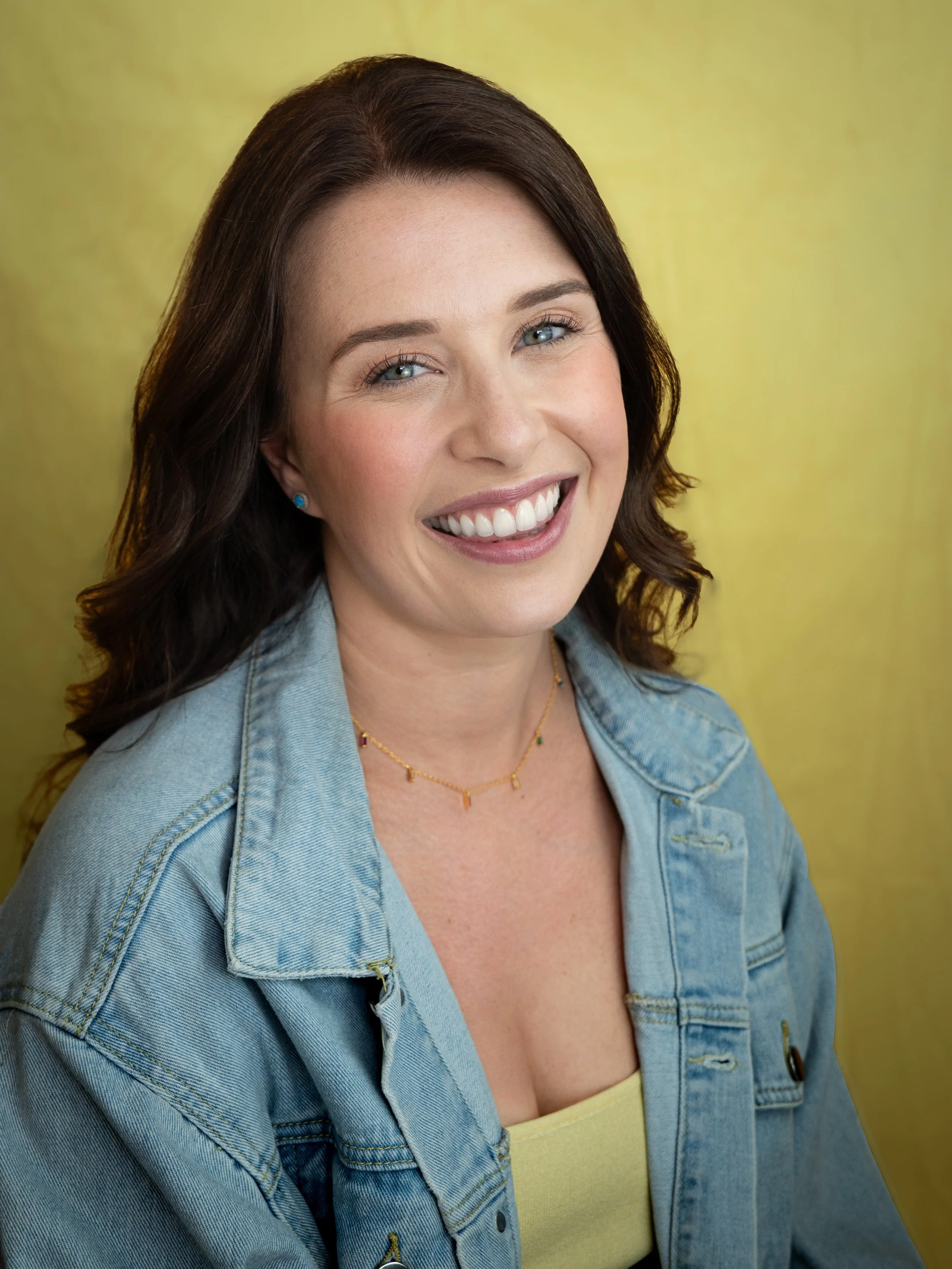 An actors headshot of a woman sitting infront of a yellow background. She is smiling widely and is wearing a light blue denim jacket and a yellow top underneath. She has dark brown hair and blue eyes. She is facing the camera.
