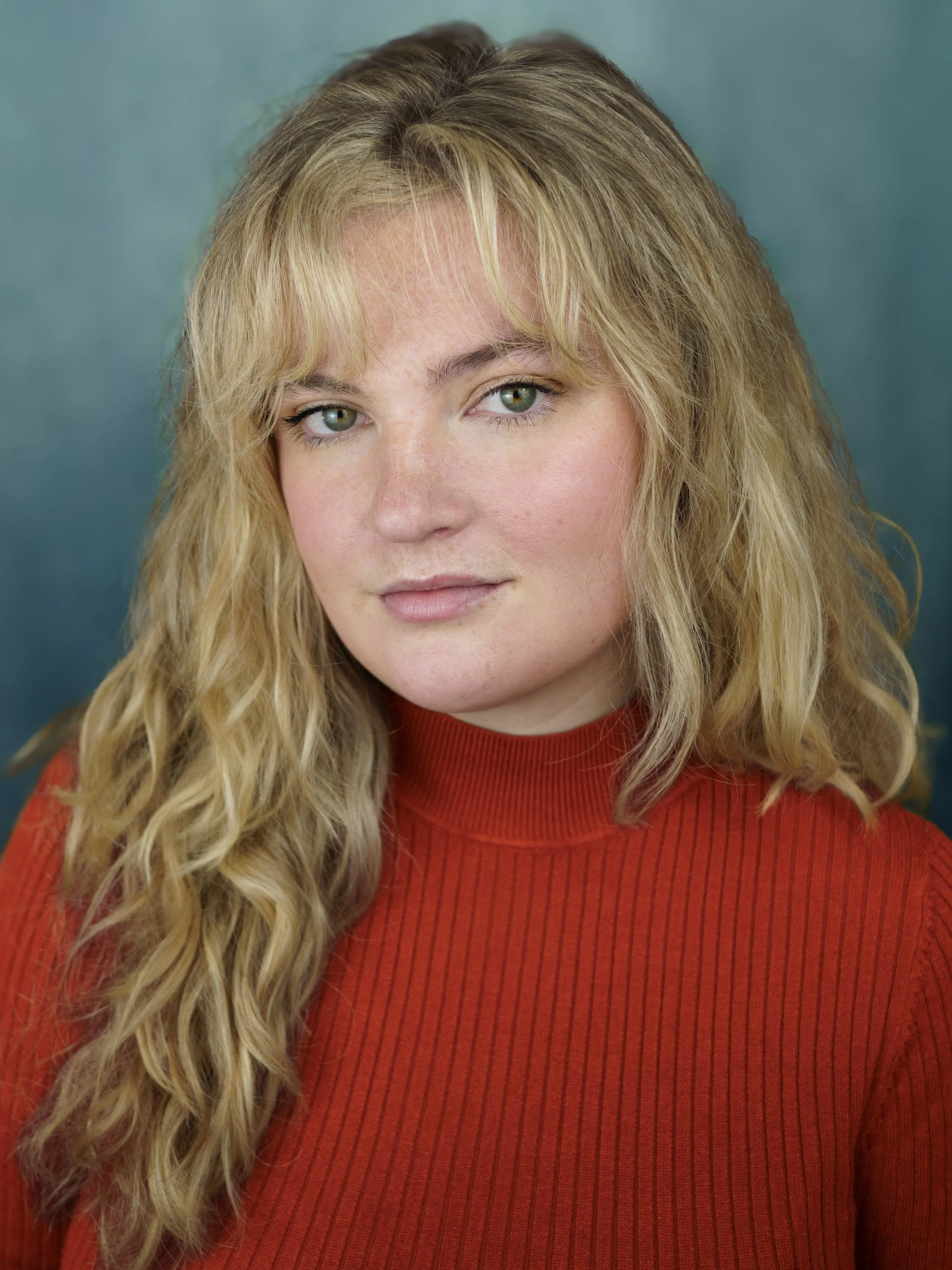An actor headshot of a woman in a rust coloured top. She has blond curly hair and a full fringe. Her eyes are green and she has a soft expression on her face. 