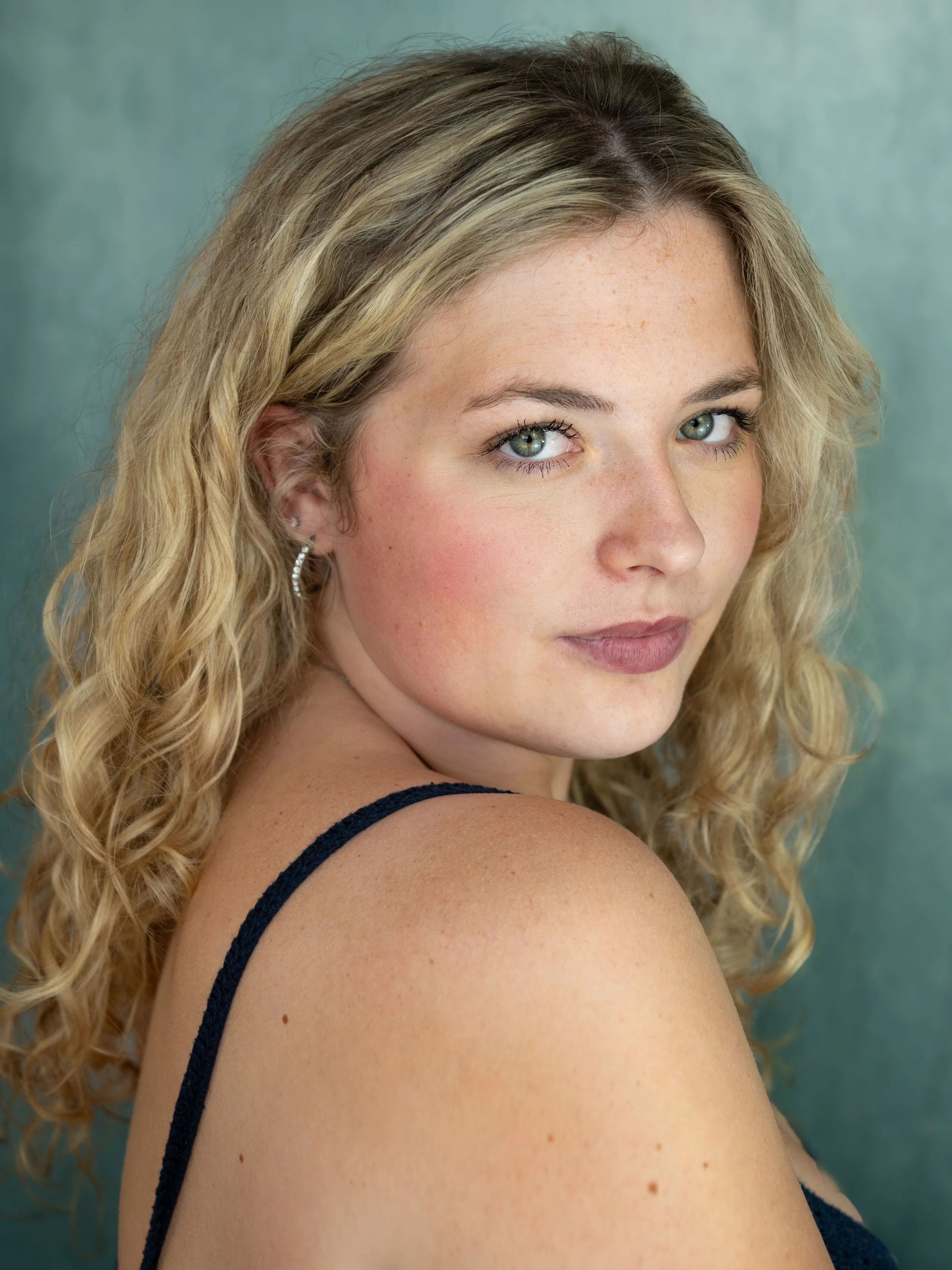 An actors headshot of a woman with blond curly hair looking over her shoulder. She has green eyes and a soft expression.