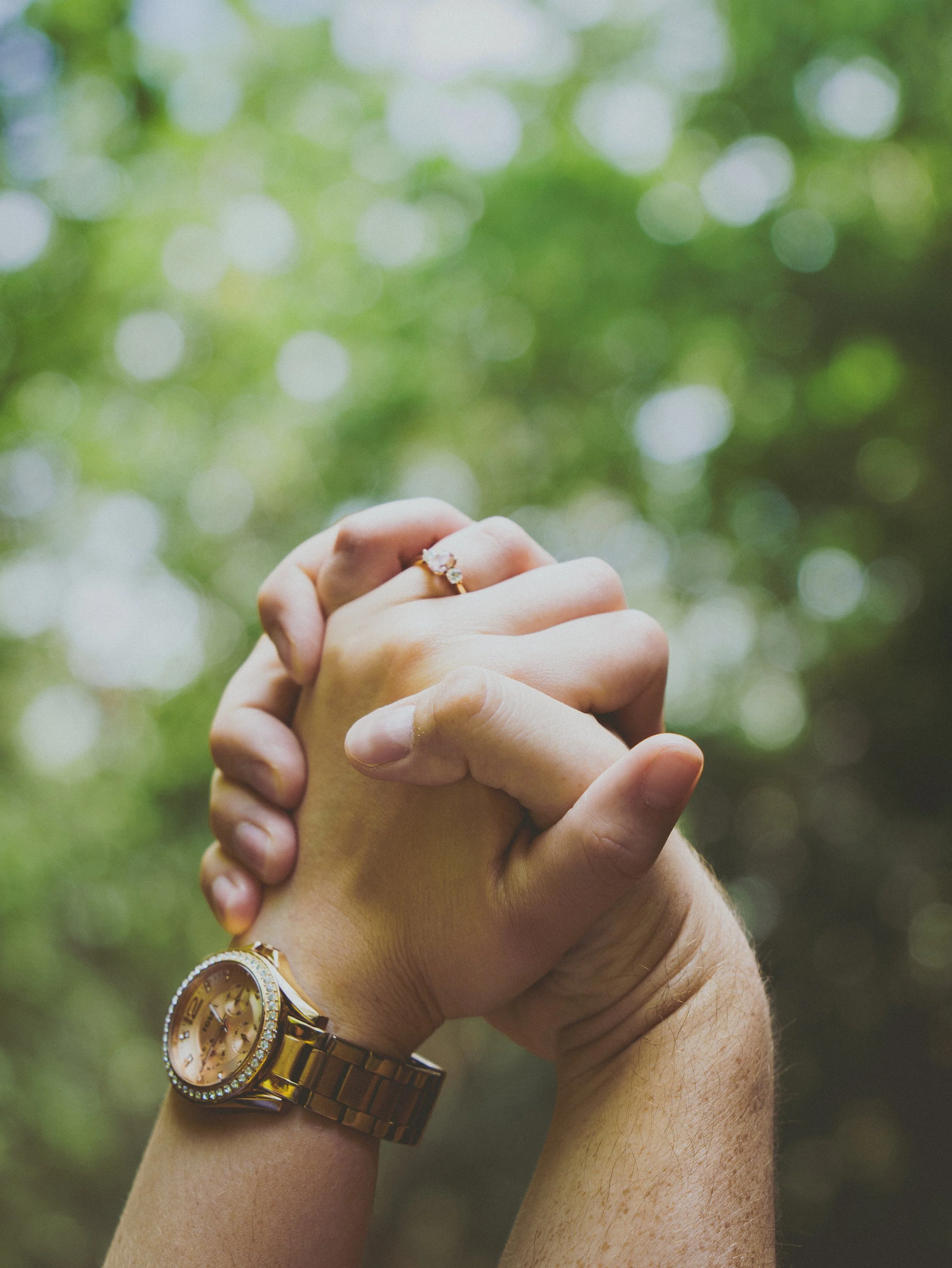 A close-up of a couple holding hands intertwined, showing an engagement ring on the woman's finger and a gold watch on the man's wrist, with a blurred green outdoor background.
