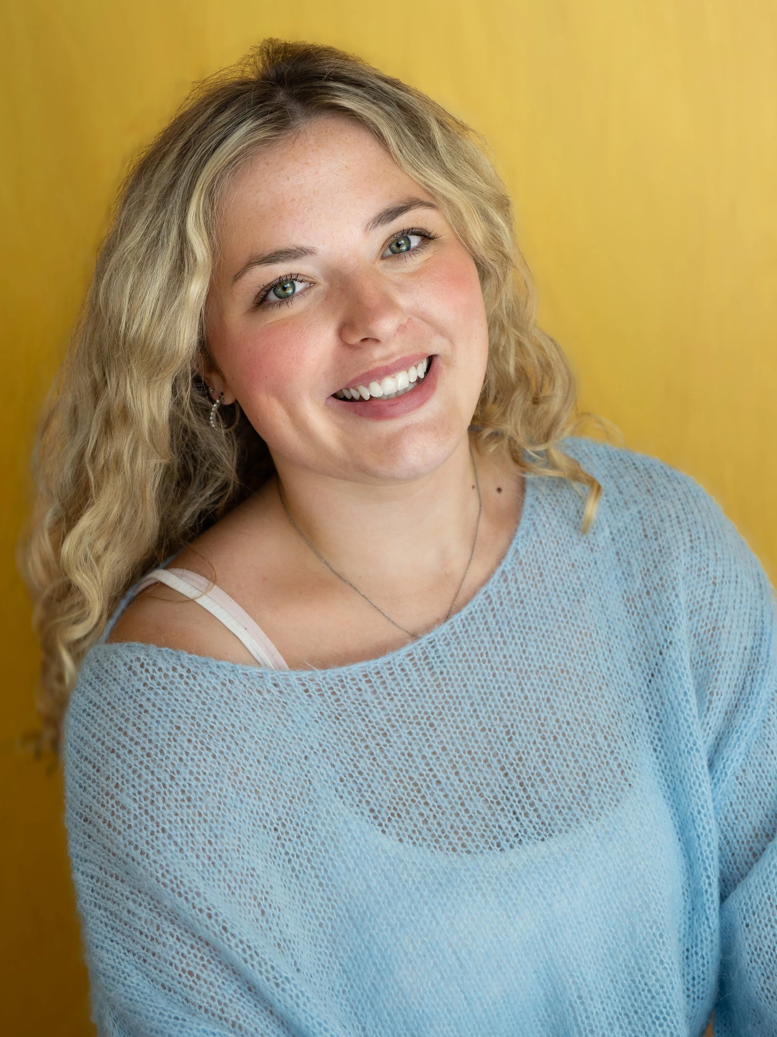 An actors headshot of a woman sitting infront of a bright yellow background. She is smiling brightly. She has blond curly hair, green eyes and is wearing a blue knit off-the-shoulder top. 