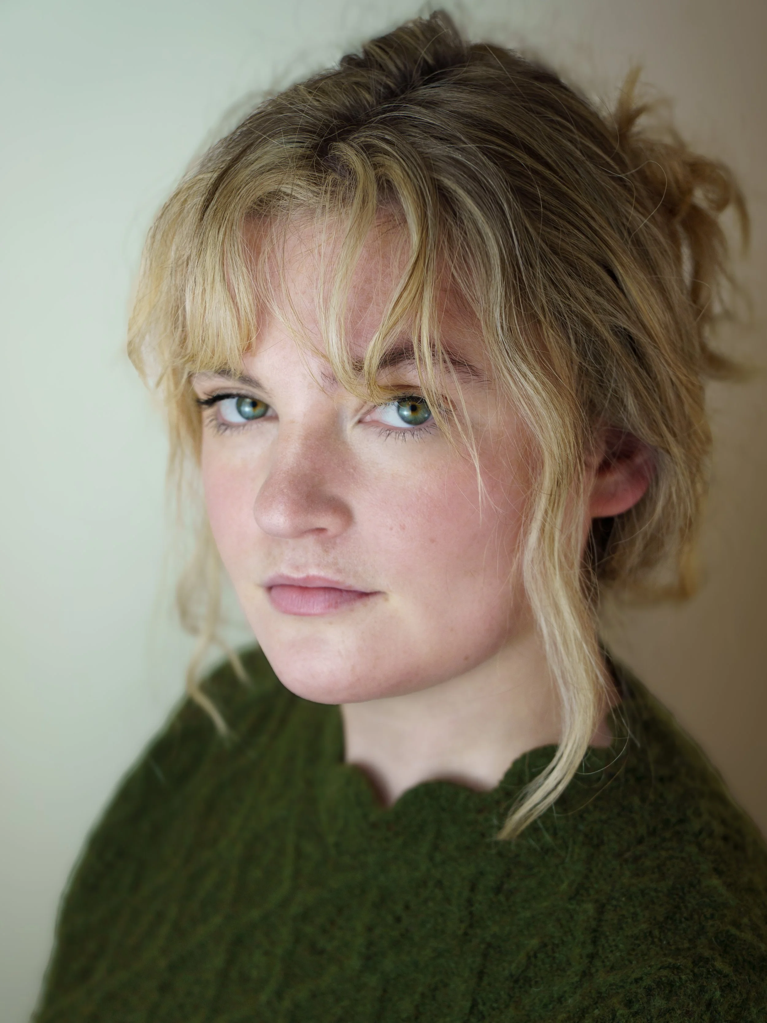 An actresses headshot on a woman side on to the camera. She has blond curly hair tied up in a bun with pieces of her fringe framing her face. She has green eyes and is wearing a green jumper. 
