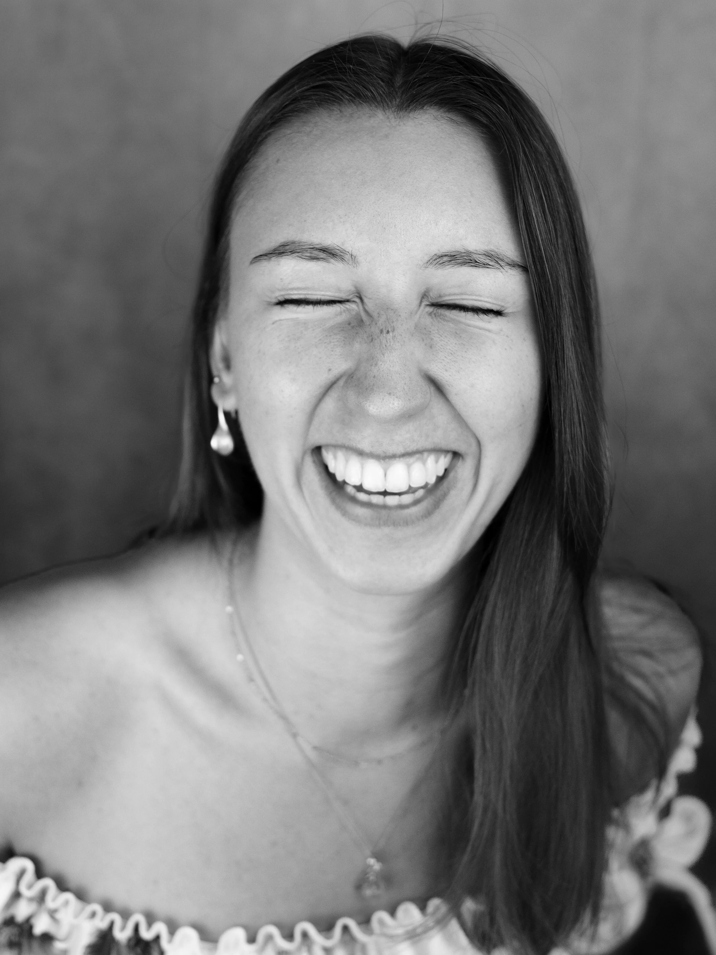 A woman with straight hair smiling widely with her eyes closed, showing her teeth, wearing earrings and a necklace, and an off-the-shoulder top, against a plain background.