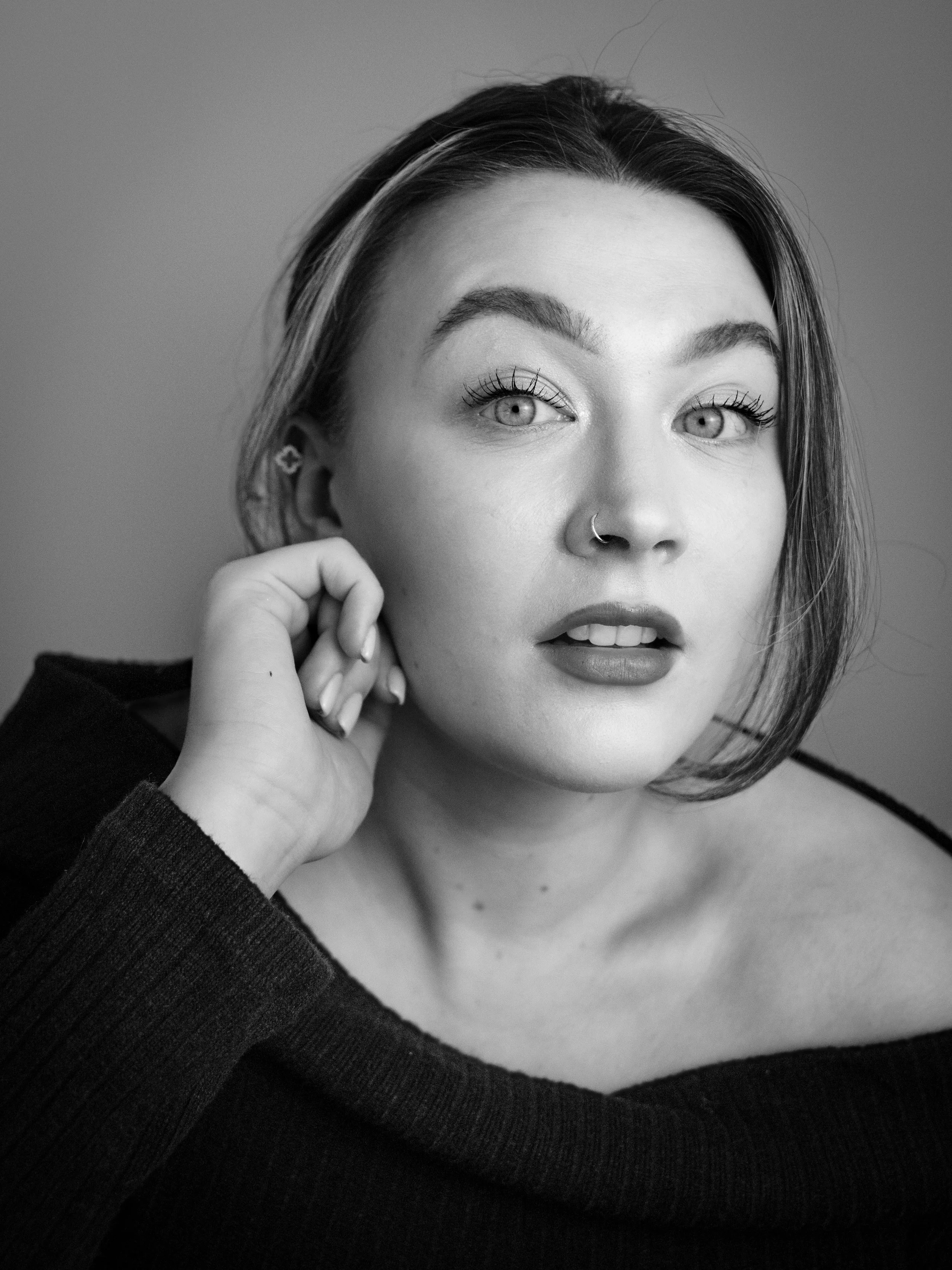 Black and white portrait of a young woman with light-colored eyes, diamond nose ring, and long eyelashes. She has dark eyebrows, shoulder-length hair, and is wearing a dark off-the-shoulder sweater. She rests her head on her left hand with fingers ne