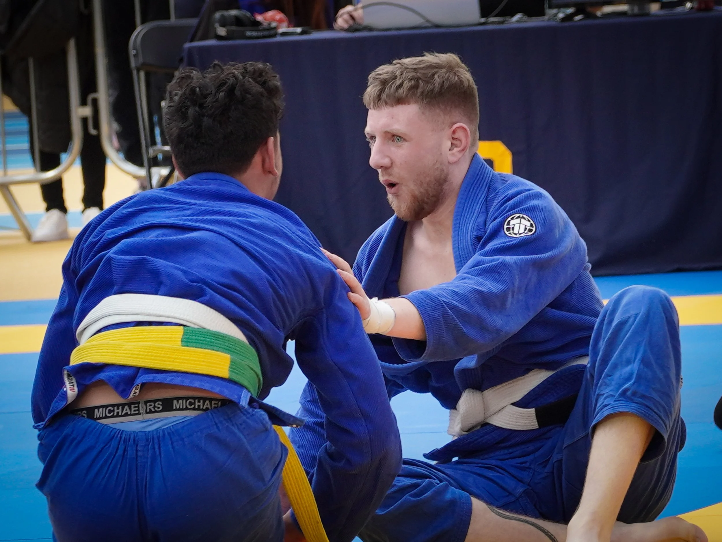 Two men in blue Brazilian Jiu-Jitsu gis sparring on a blue mat, one sitting on the ground and the other kneeling, engaging in grappling practice during a competition or training session.