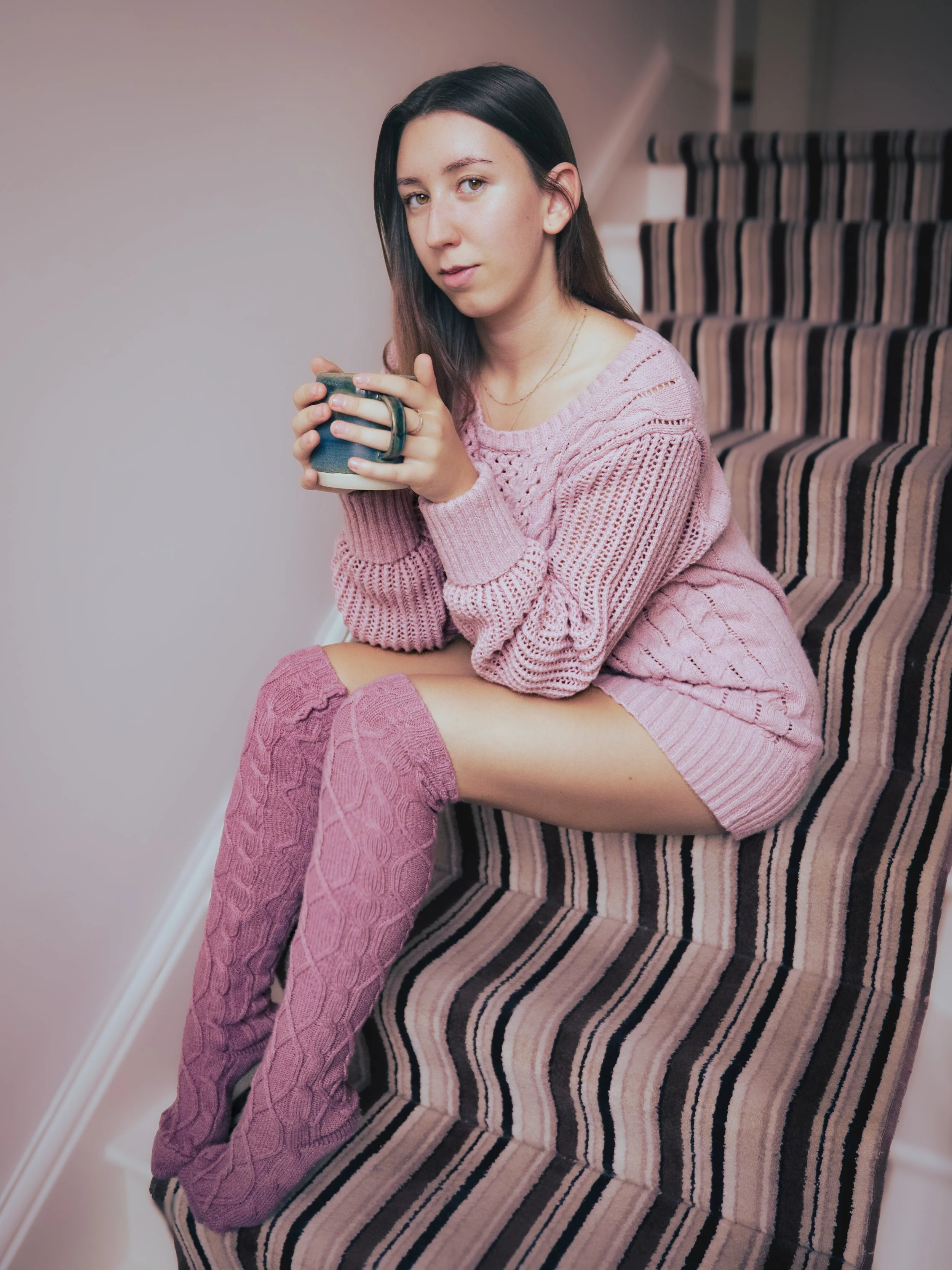 A young woman with dark hair and fair skin sitting on a staircase with striped carpet, wearing a pink knitted sweater and thigh-high pink cable knit socks, holding a mug and looking at the camera.